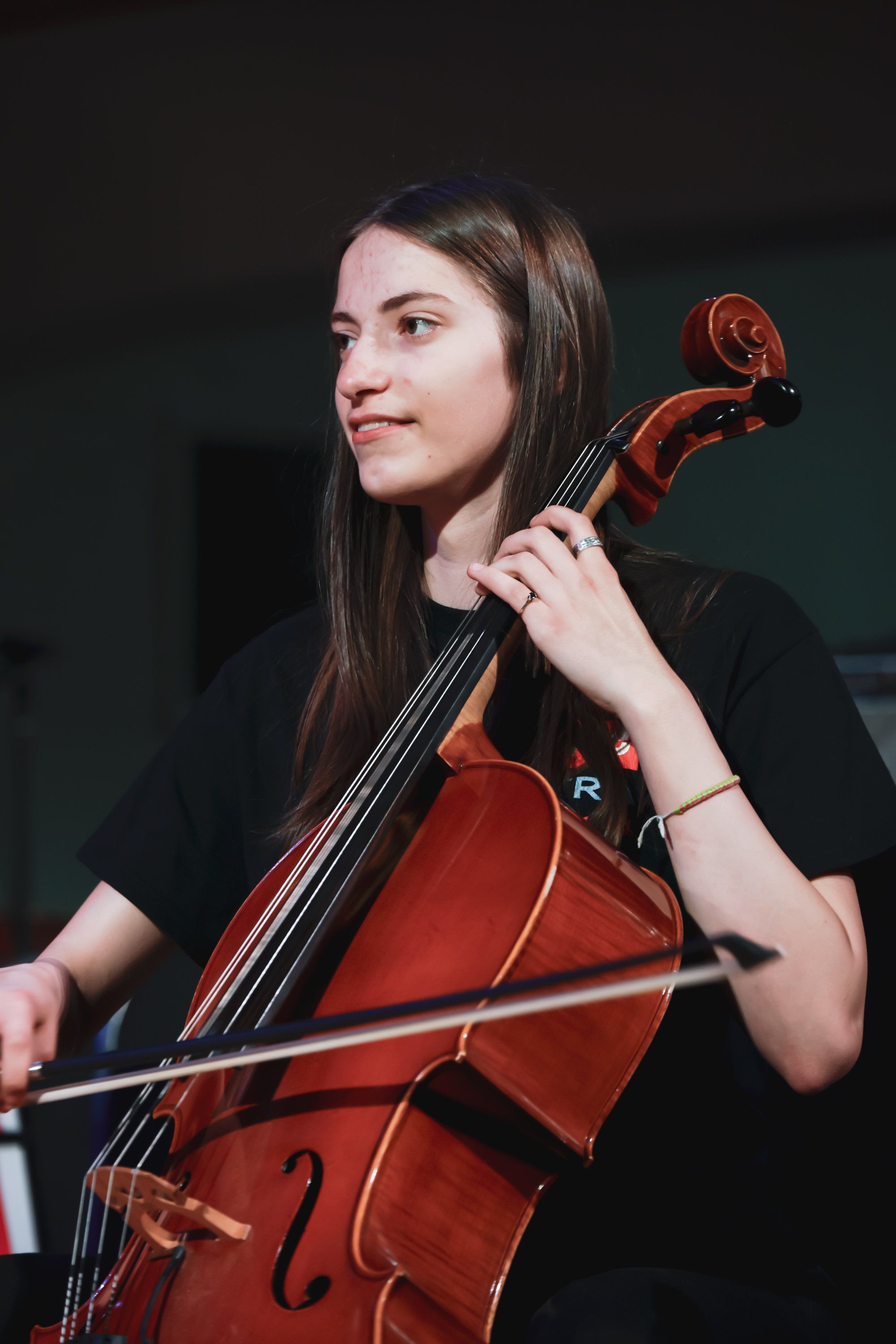 Cellist in black shirt holds instrument and bow, poised mid-performance against dark background with warm stage lighting