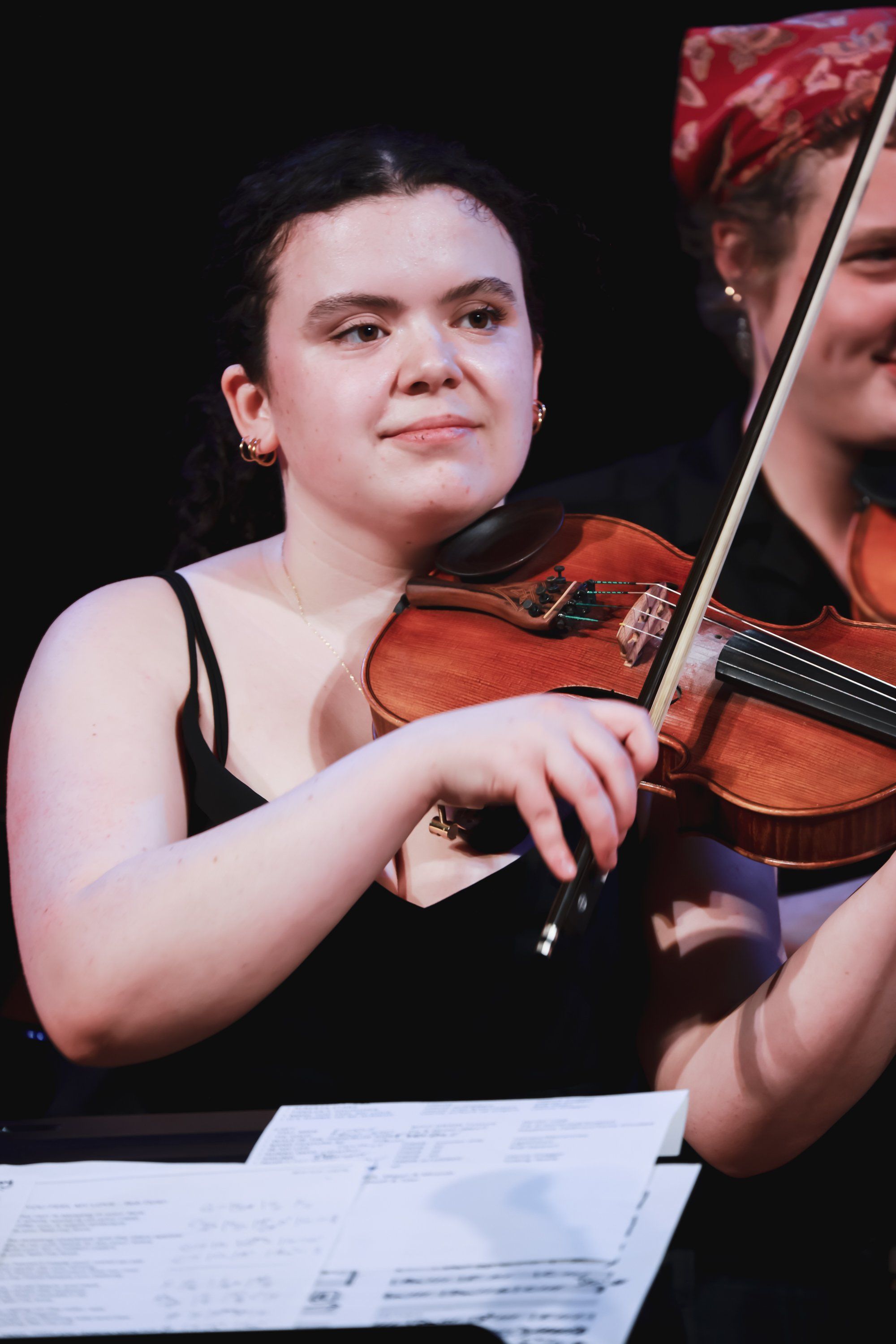 Young musician in black sleeveless top playing violin against dark background, sheet music visible below