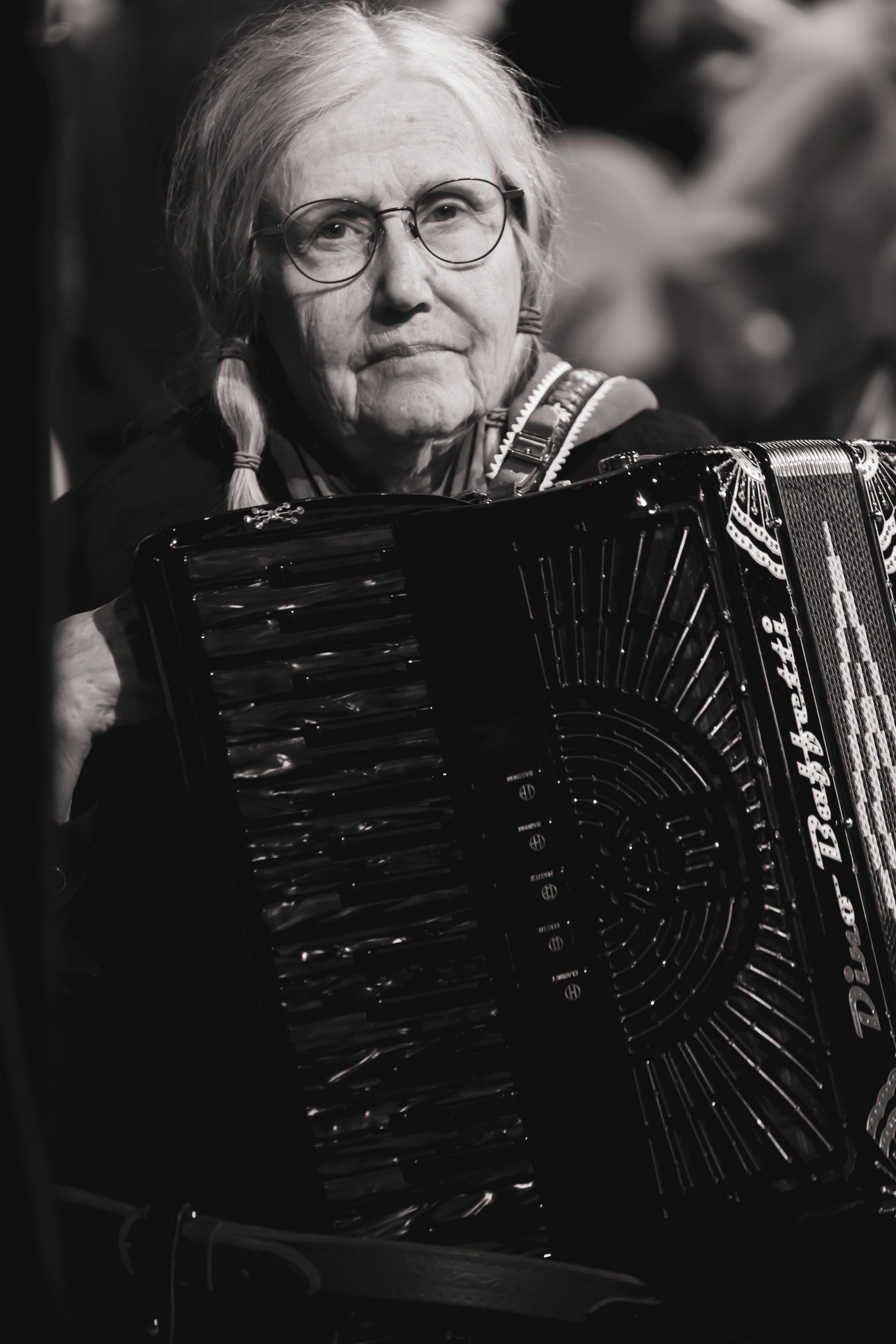 Elderly musician with white hair and glasses holds an accordion, captured in dramatic black-and-white portrait lighting