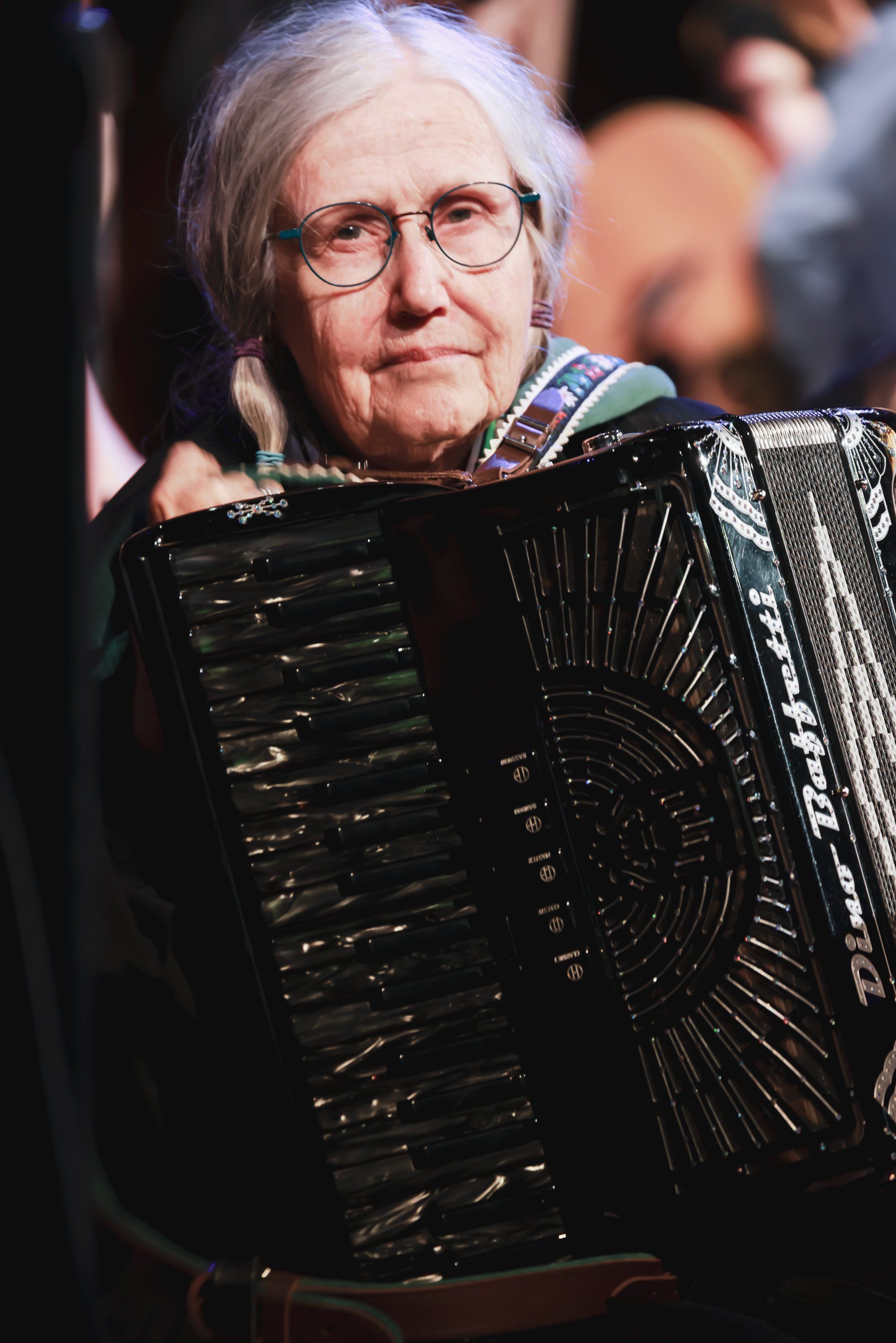 Older man wearing glasses and white hair plays a large black accordion on stage, warmly lit against a softly blurred background