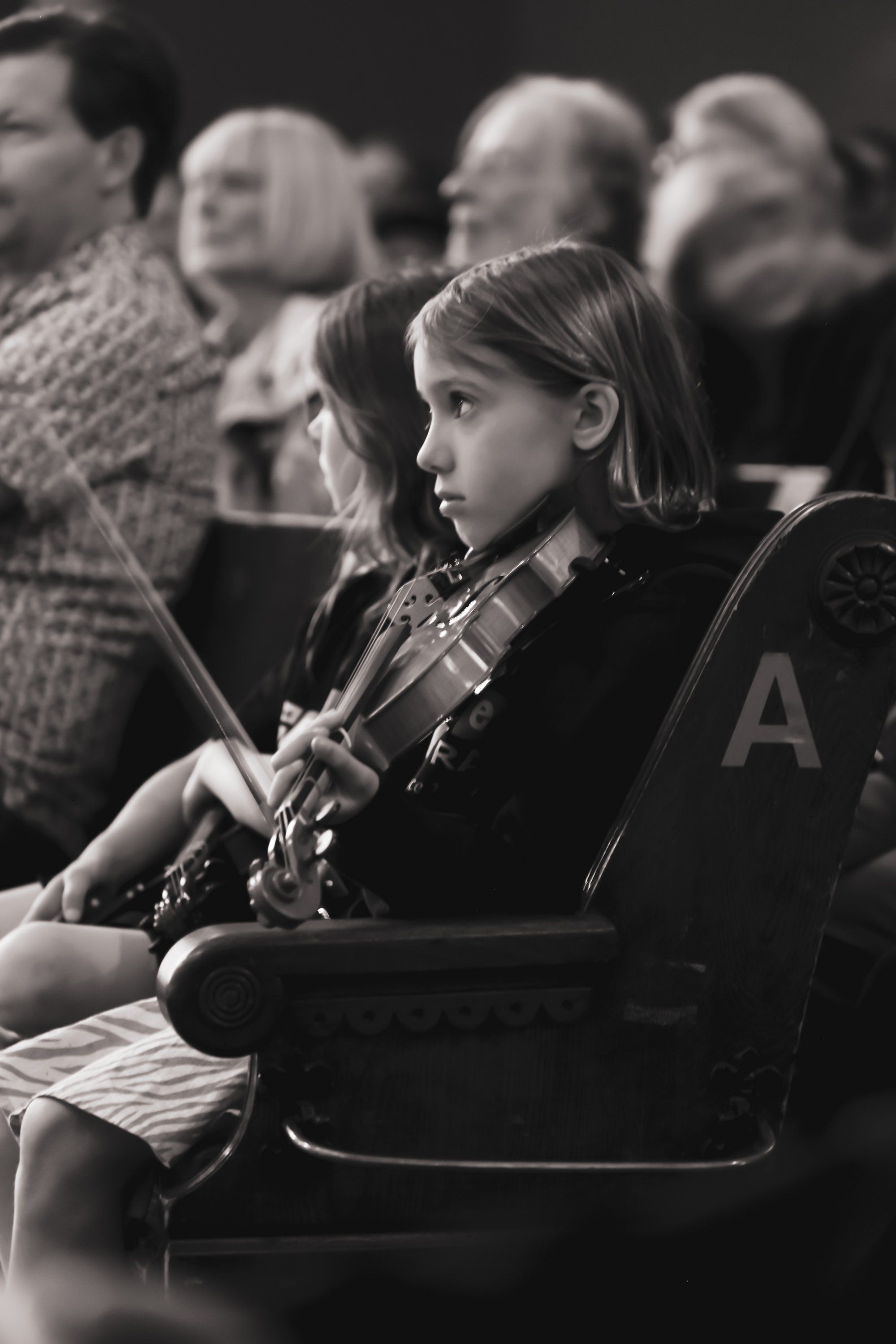 Young violinist in formal attire concentrates intently while performing, with blurred audience members seated behind in black and white