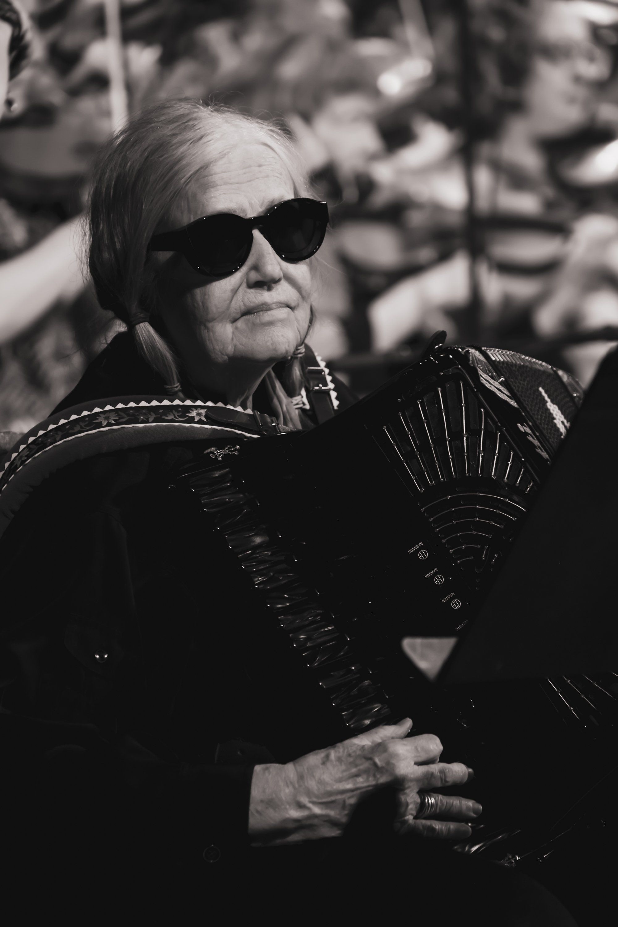 Older man in dark sunglasses and ornate black jacket holds accordion, contemplative expression against blurred outdoor background