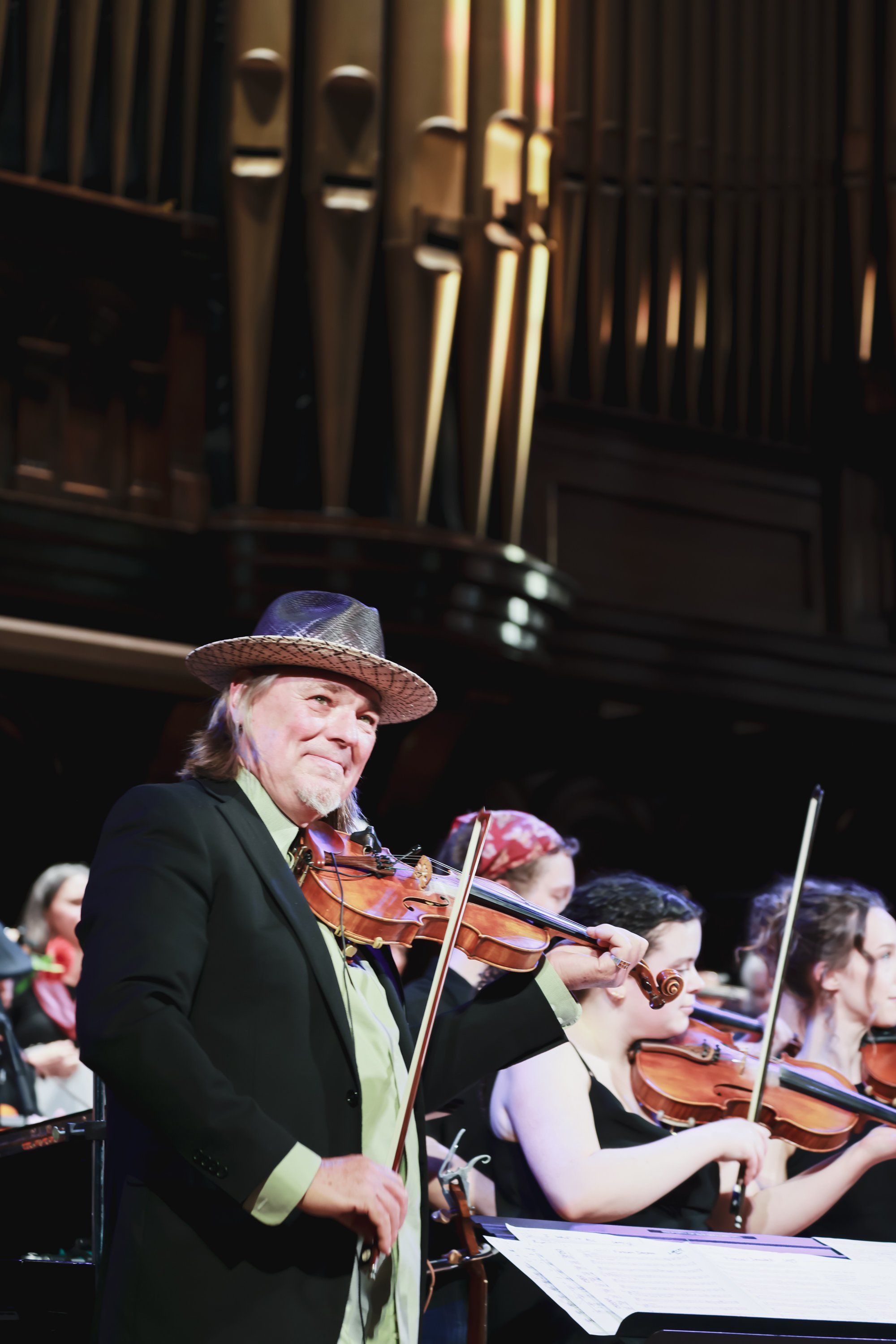 Violinist in cowboy hat and green vest performs solo under warm stage lights with orchestra members seated behind him
