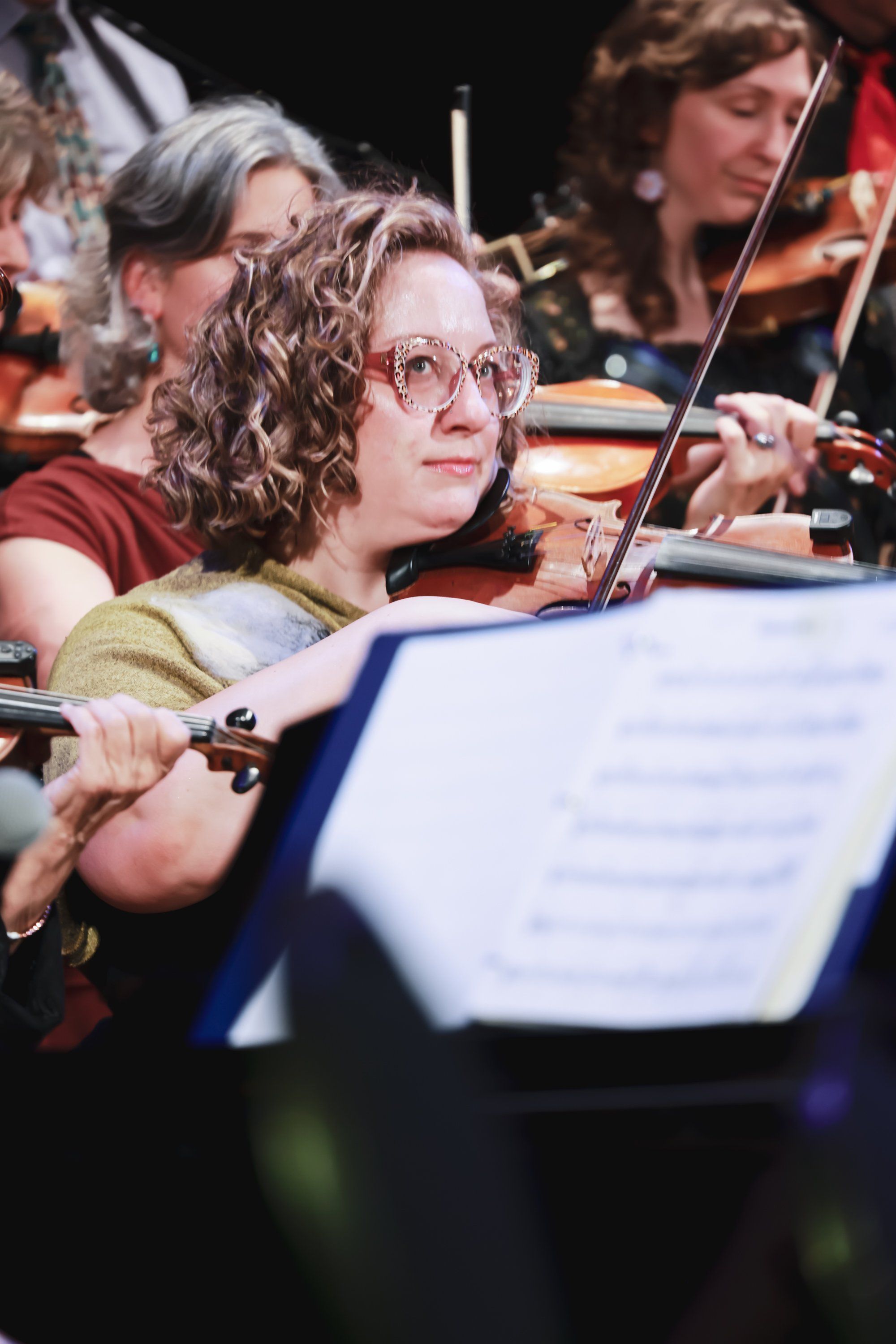 Violinist in glasses and burgundy shirt focuses intently on sheet music during an orchestral performance, with ensemble members visible in soft background li…