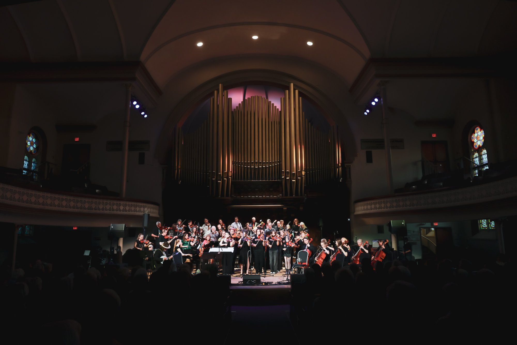 Choir and orchestra performing on stage beneath a grand pipe organ in a warmly lit church sanctuary