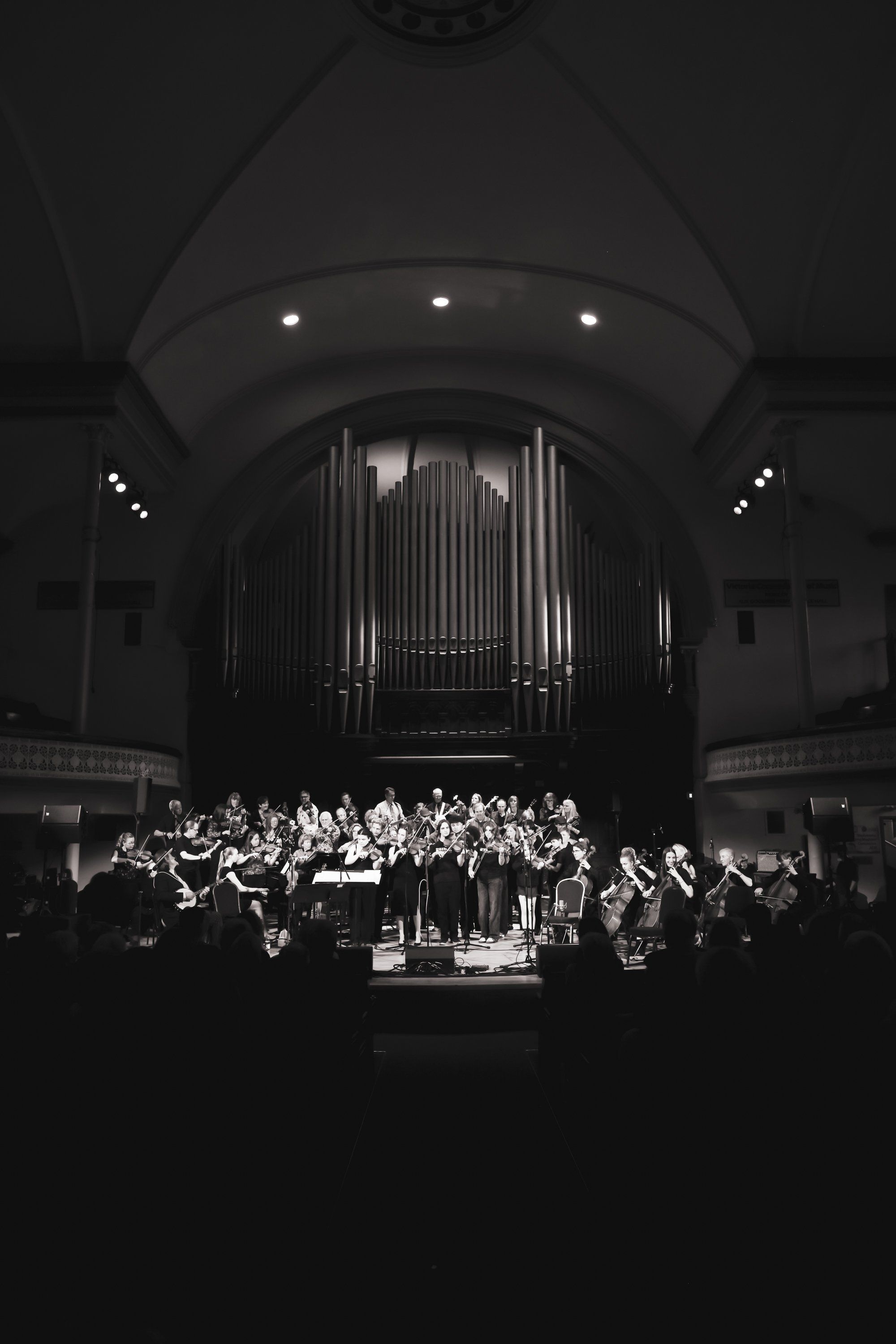 Orchestra and choir perform beneath a grand pipe organ in a dramatically lit concert hall, captured in black and white