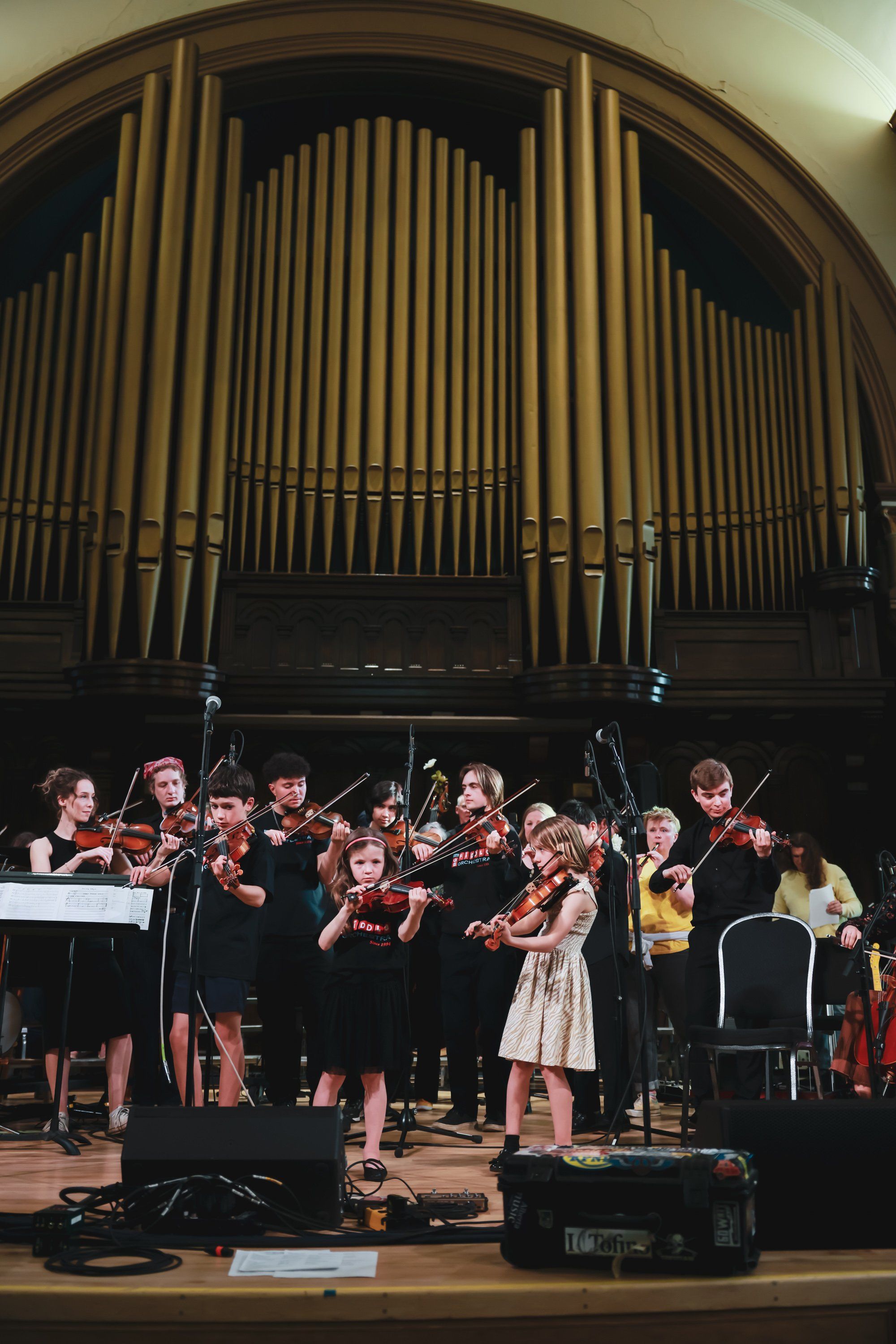 Young musicians perform with string instruments on stage beneath a grand pipe organ, bathed in warm spotlight