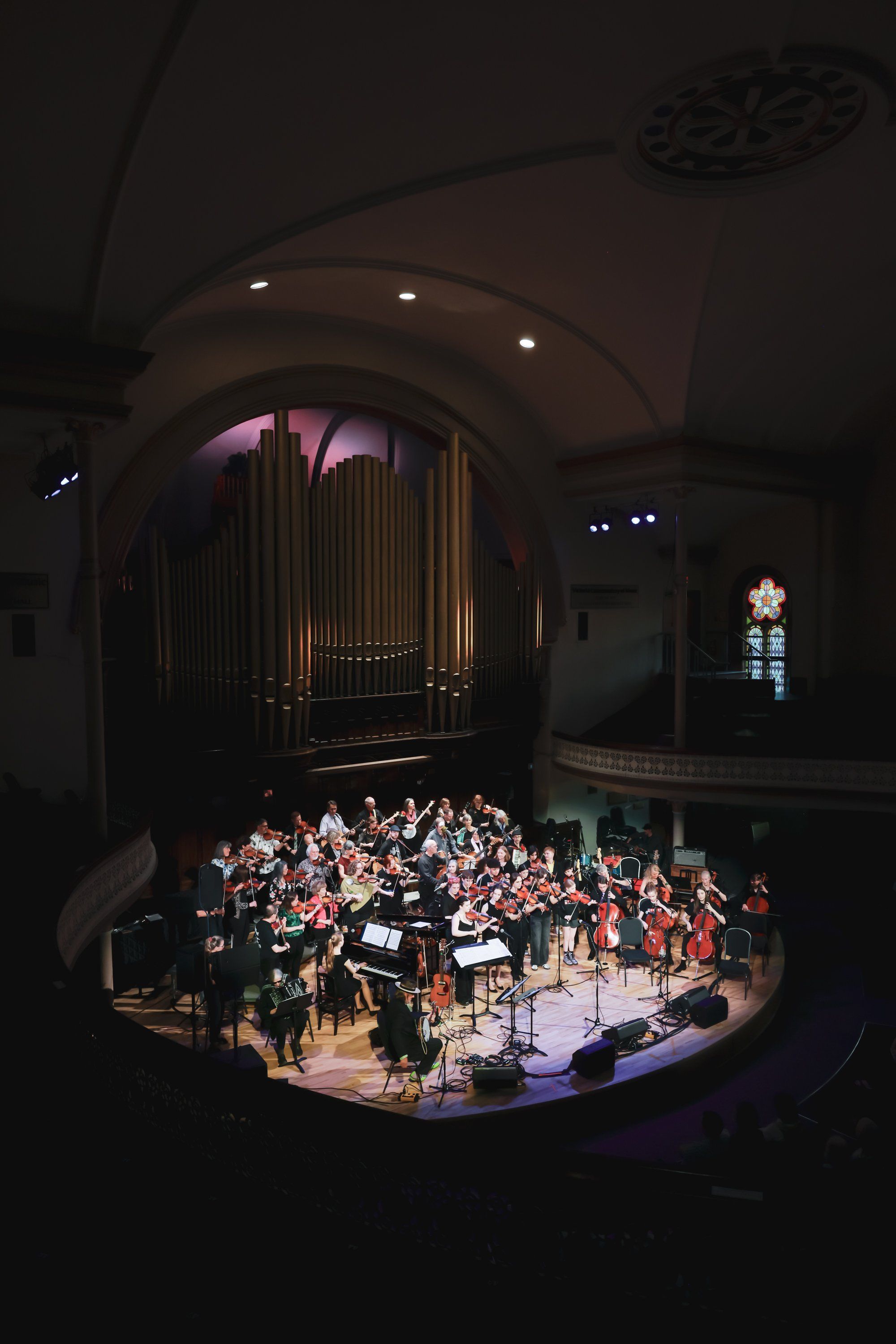 Orchestra and conductor illuminated on concert hall stage beneath a grand pipe organ, audience seated in warm spotlight