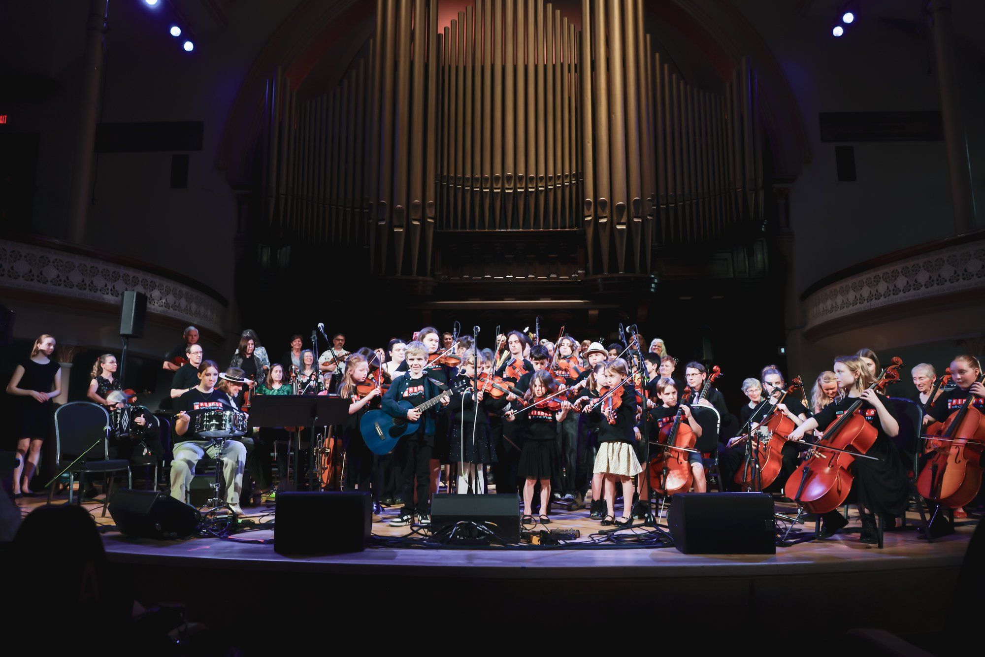 Large orchestra and choir performing on stage beneath a grand organ, bathed in warm stage lighting with dramatic shadows