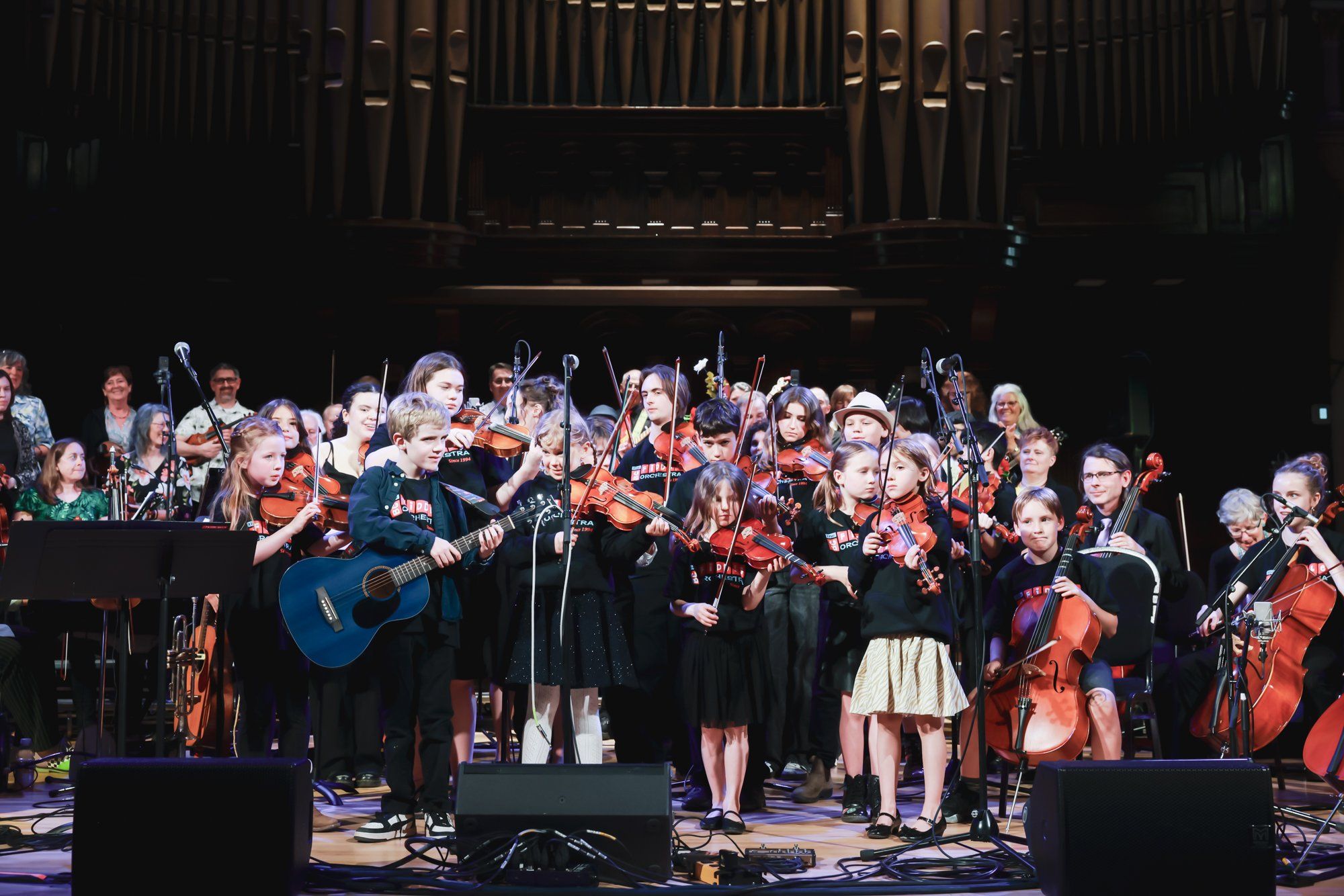 Orchestra and choir members perform together on stage under warm lighting, with string instruments and acoustic guitar prominent in the foreground