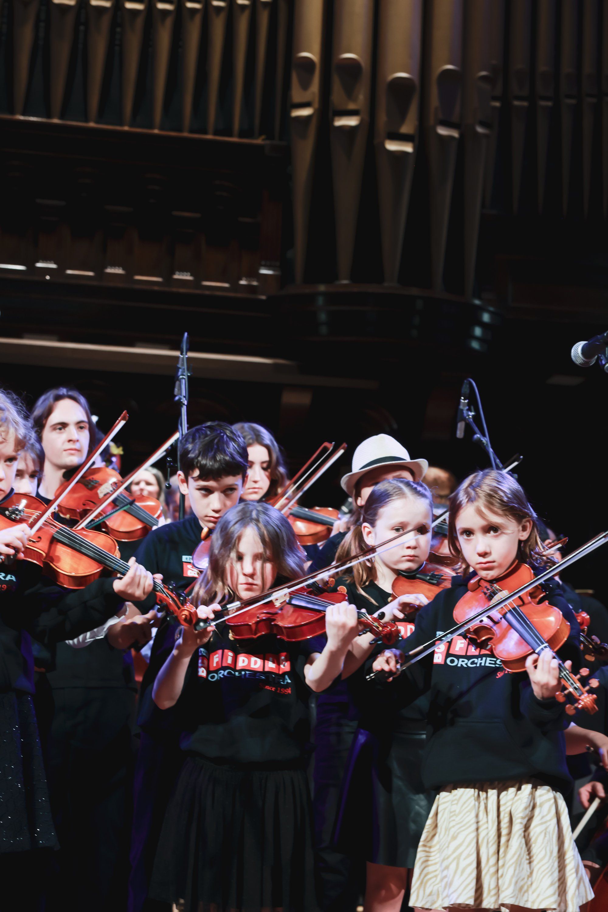 Young violinists in dark uniforms perform together on stage, bathed in warm light with a dark organ backdrop
