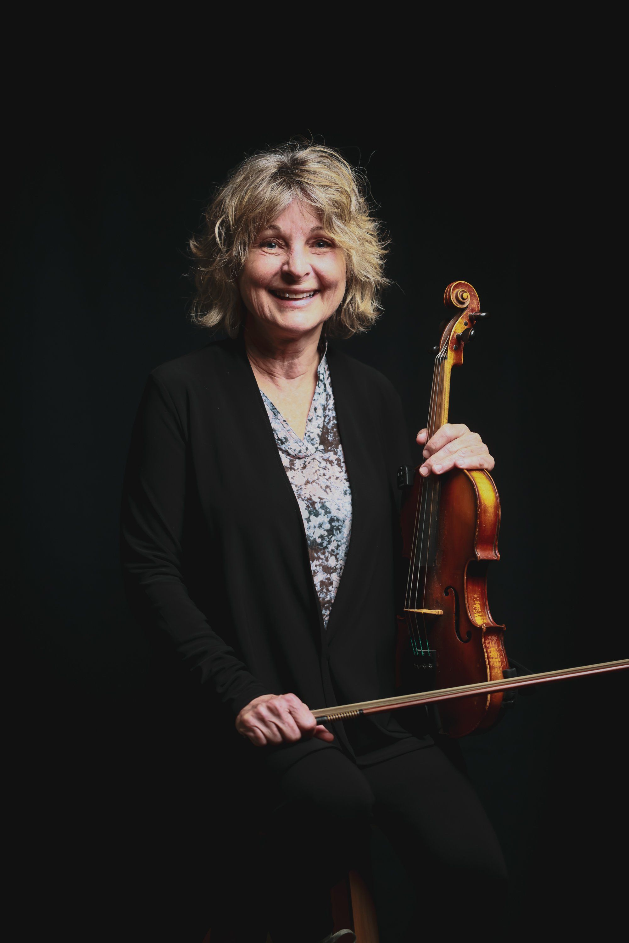 Cellist with blonde wavy hair in black blazer holds instrument and bow against dark background, smiling warmly at camera