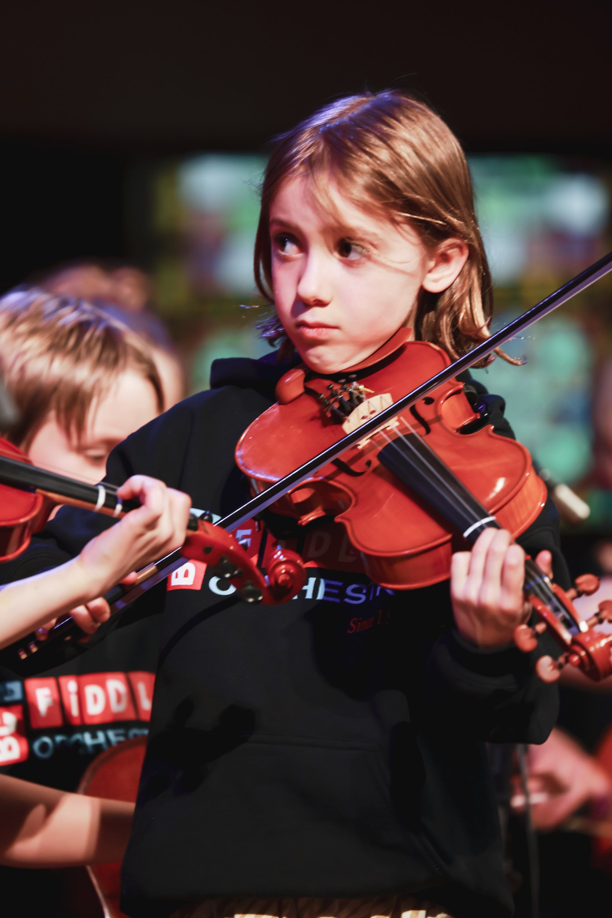 Young violinist in red instrument and dark jacket performs with focused expression during ensemble concert