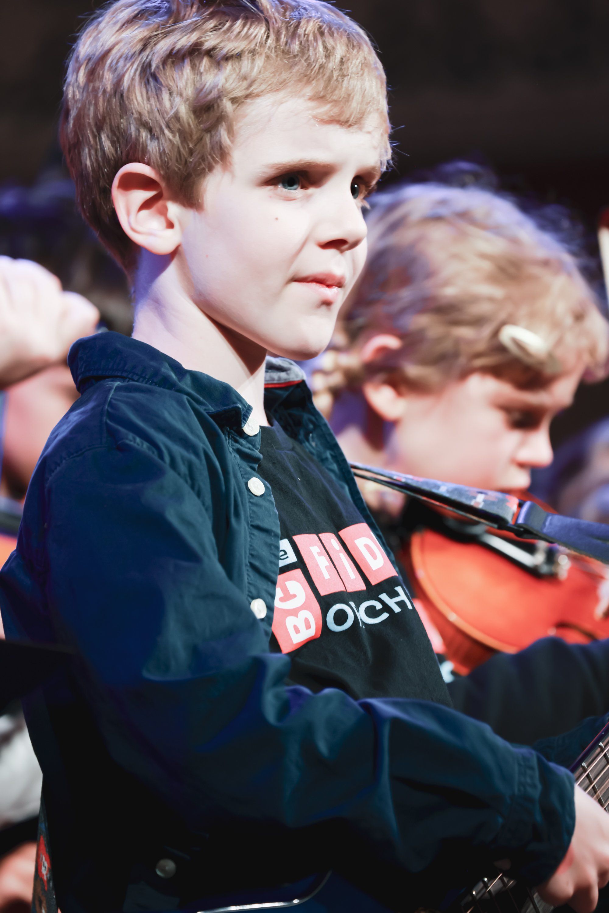 Young boy in dark jacket holding violin, focused expression during performance at concert event