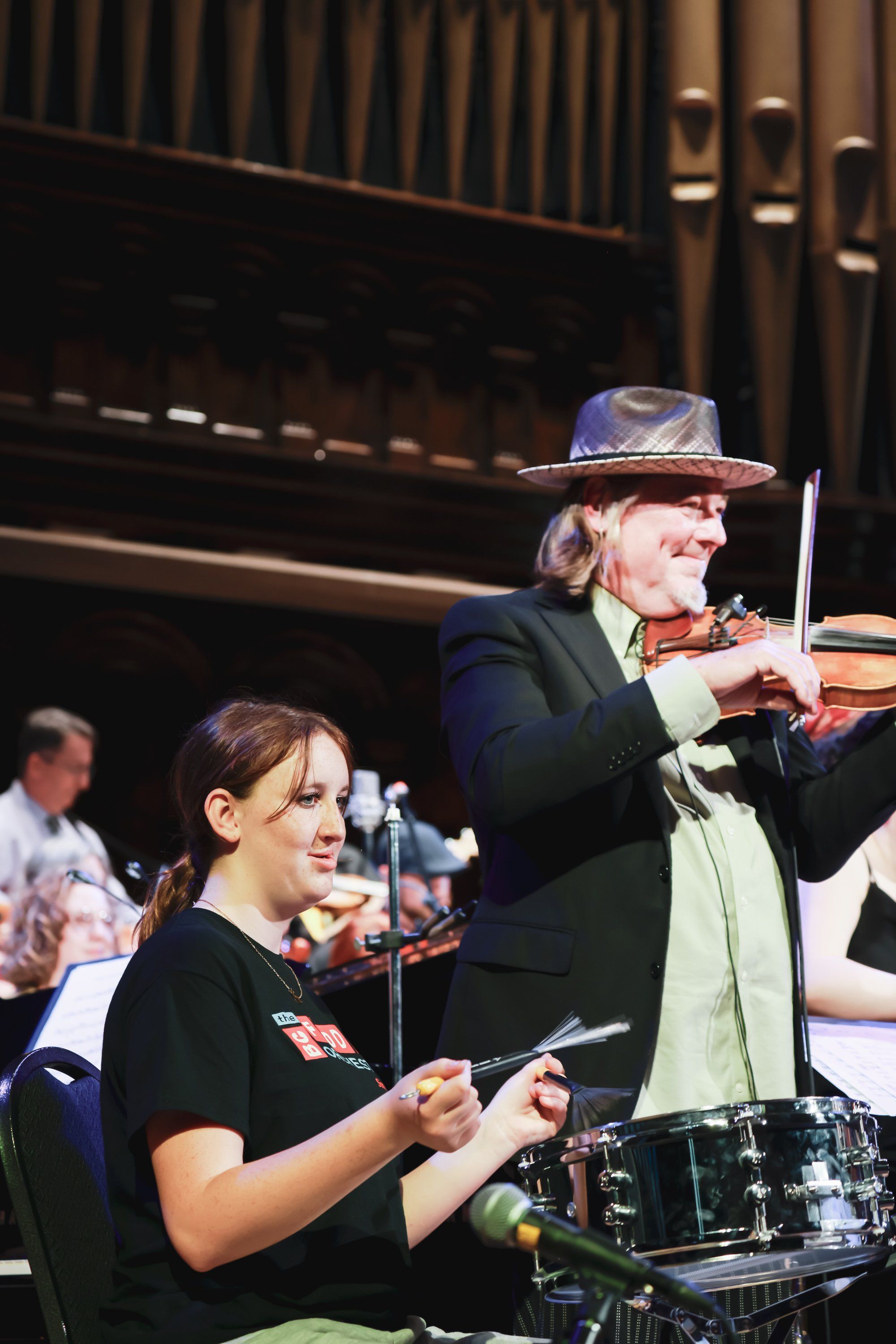 Two musicians perform at an indoor concert venue, one playing violin while wearing a cowboy hat, the other on drums, with warm stage lighting and an organ vi…