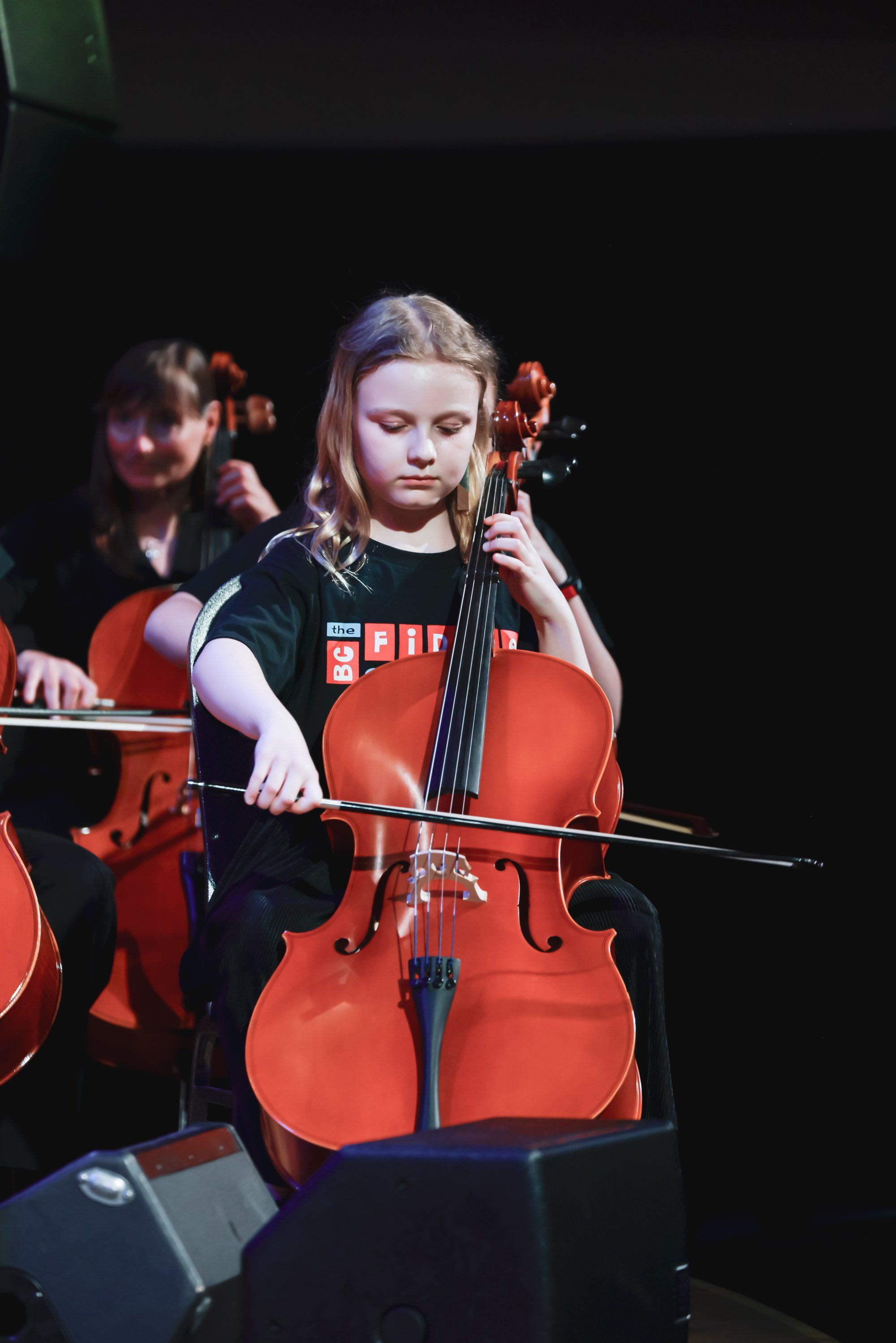 Cellist in red instrument performs on dark stage, focused expression, warm stage lighting illuminating her face and orchestra members behind her