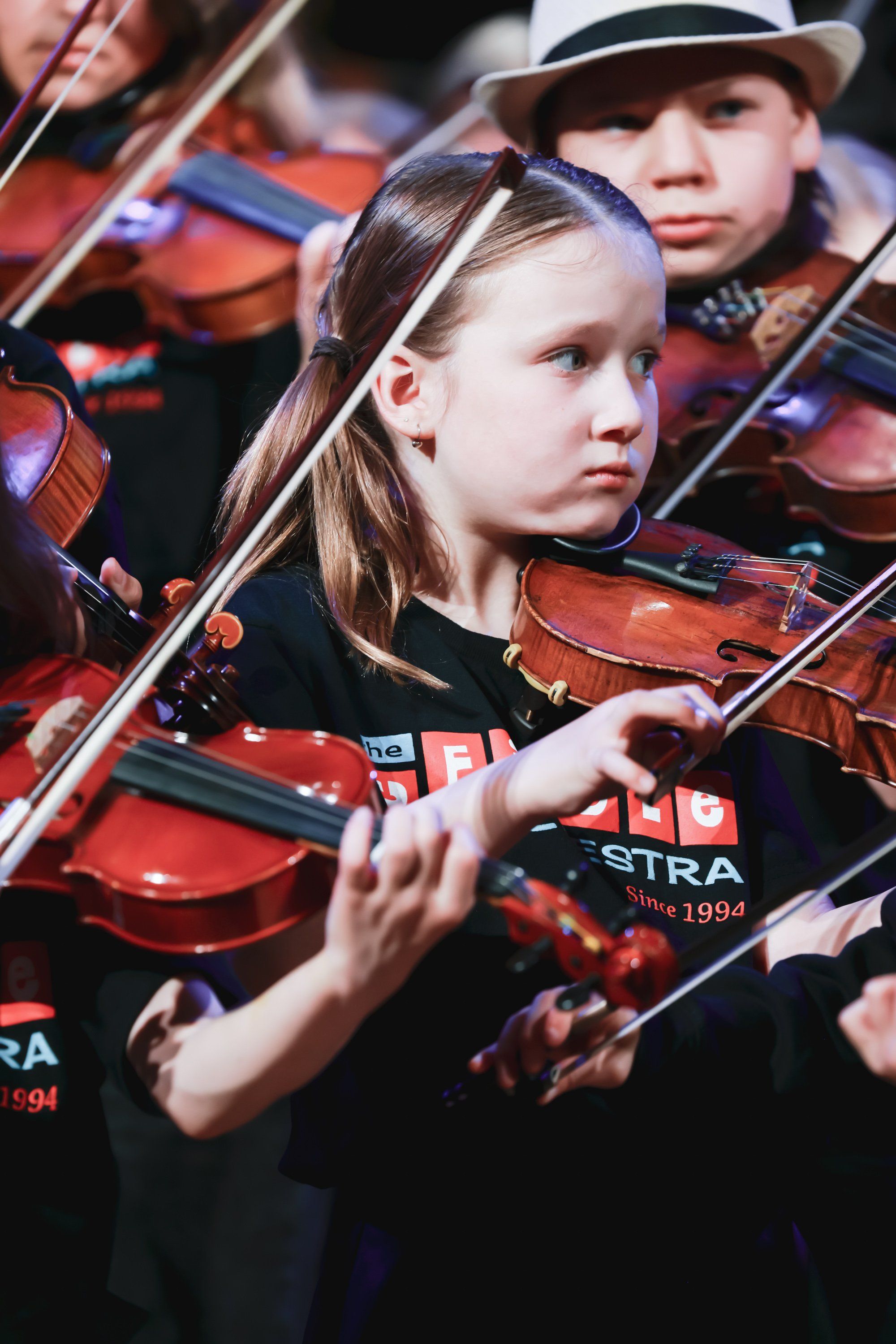 Young musicians in concert attire concentrate while playing violins, warm stage lighting reflecting off polished instruments