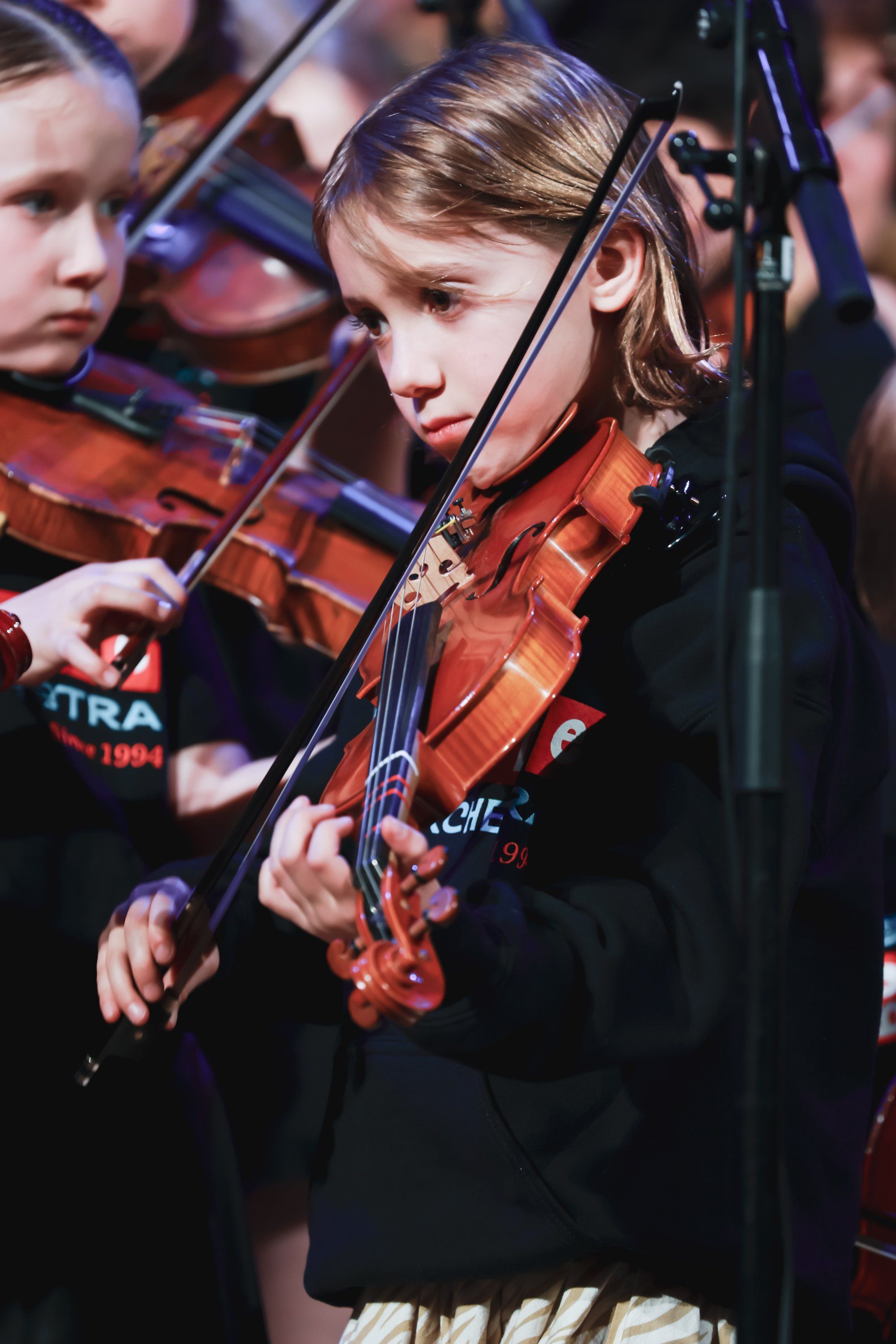 Young violinist in red instrument and black outfit performs with ensemble, warm stage lighting highlighting concentrated expression