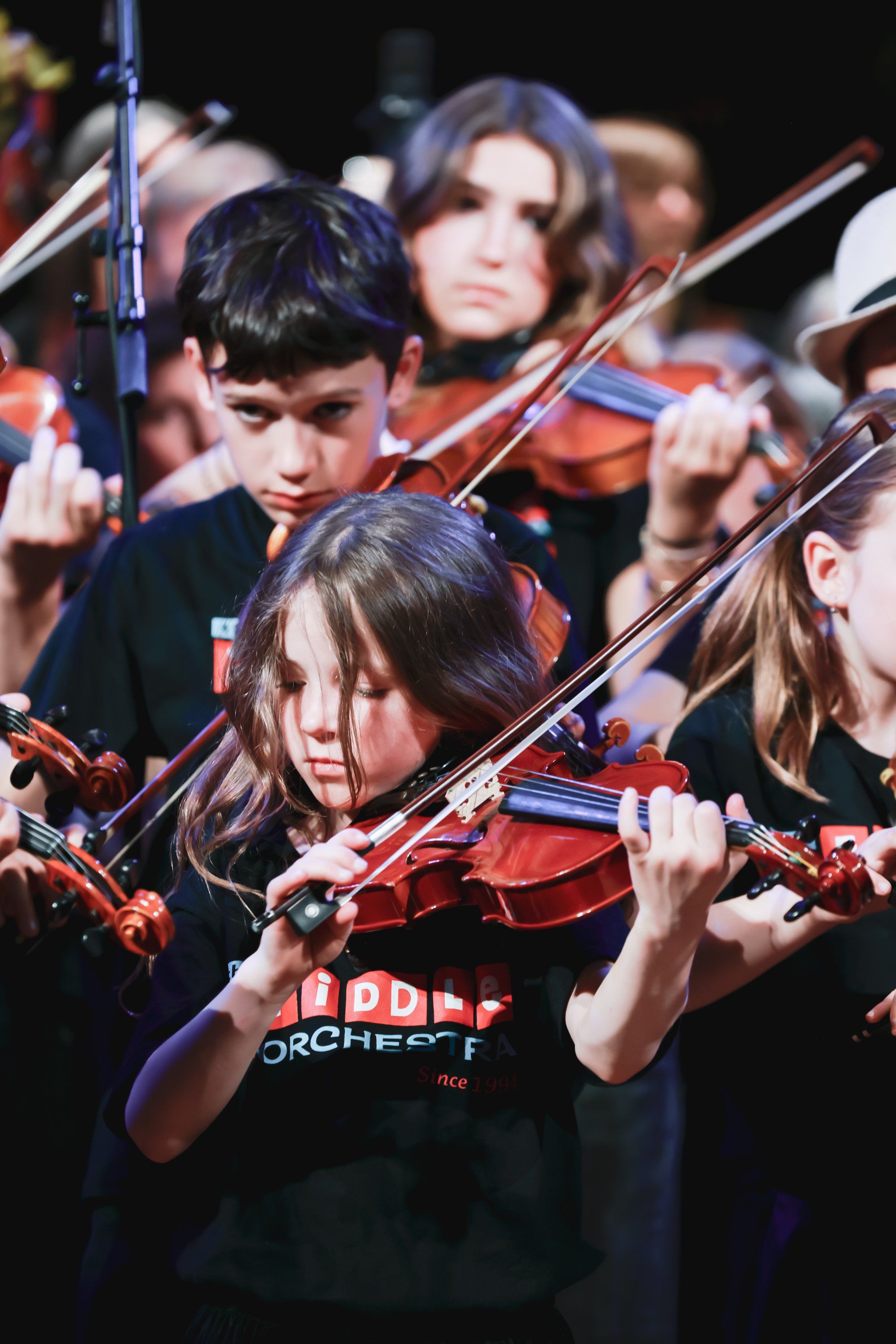 Young musicians in dark orchestra attire concentrate while playing violins together on stage under warm lighting
