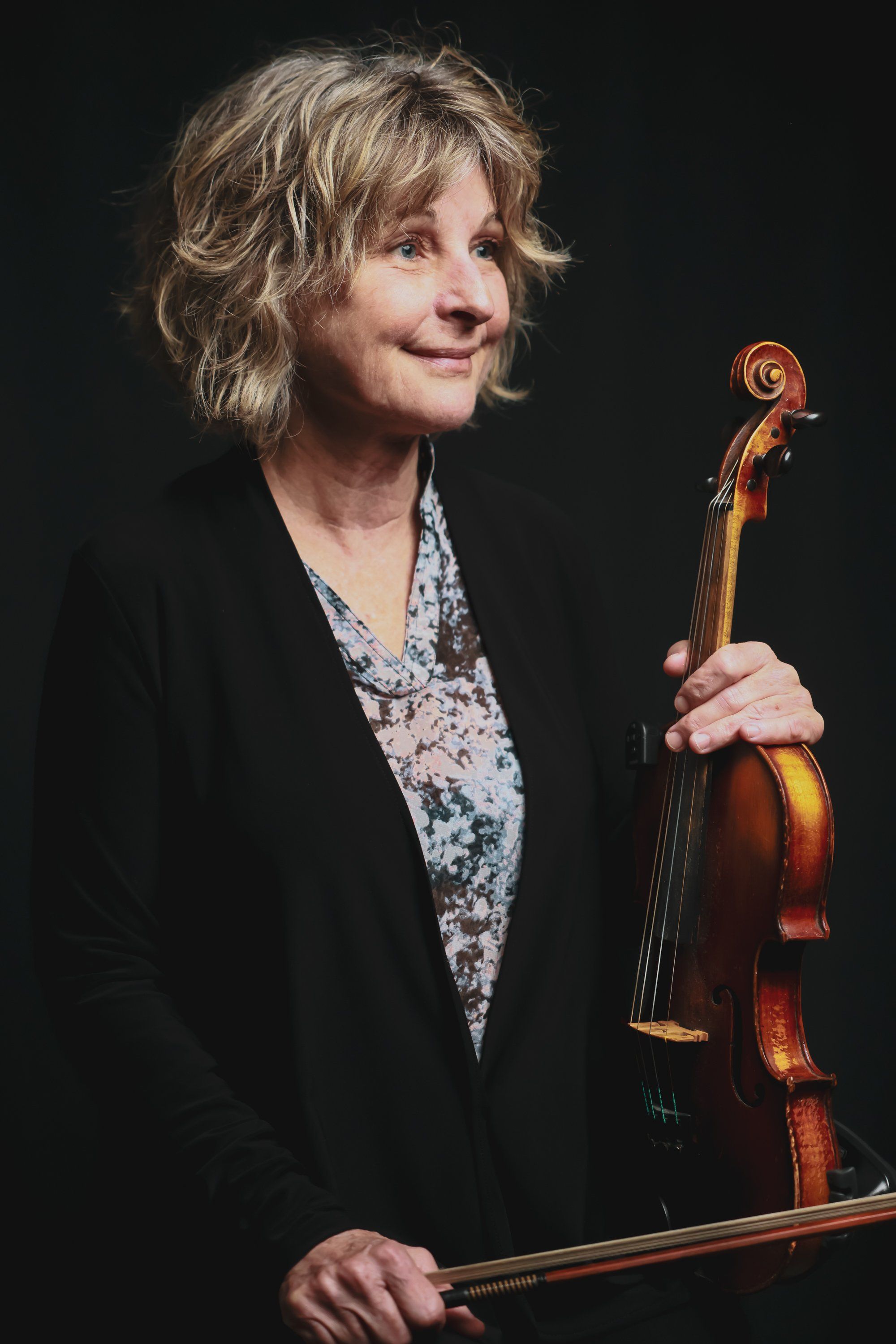 Violinist with short blonde hair holds her instrument and bow against a dark background, wearing a black blazer over a patterned blouse, gazing thoughtfully…