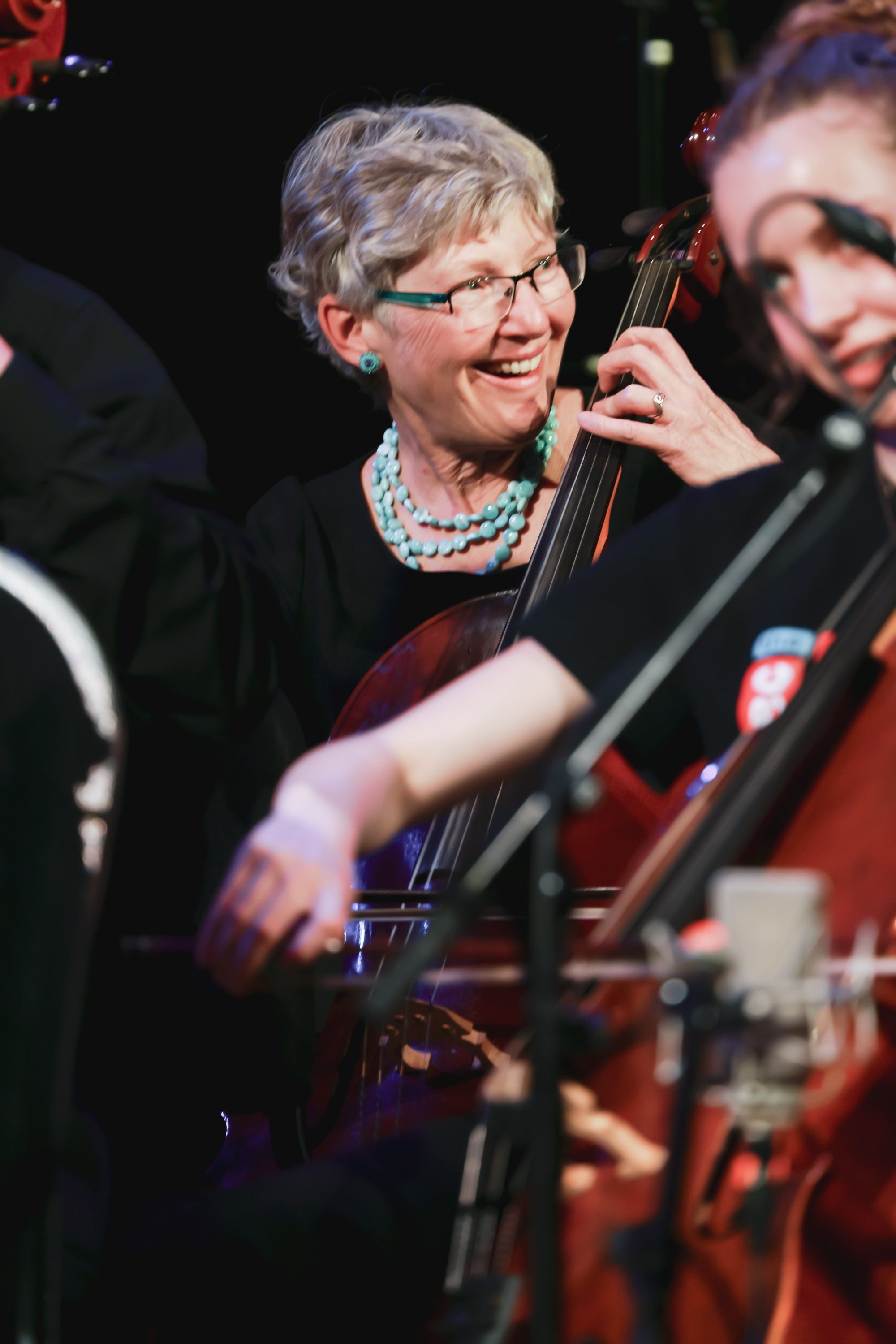 Smiling older woman with glasses playing cello among ensemble musicians on stage under warm theatrical lighting