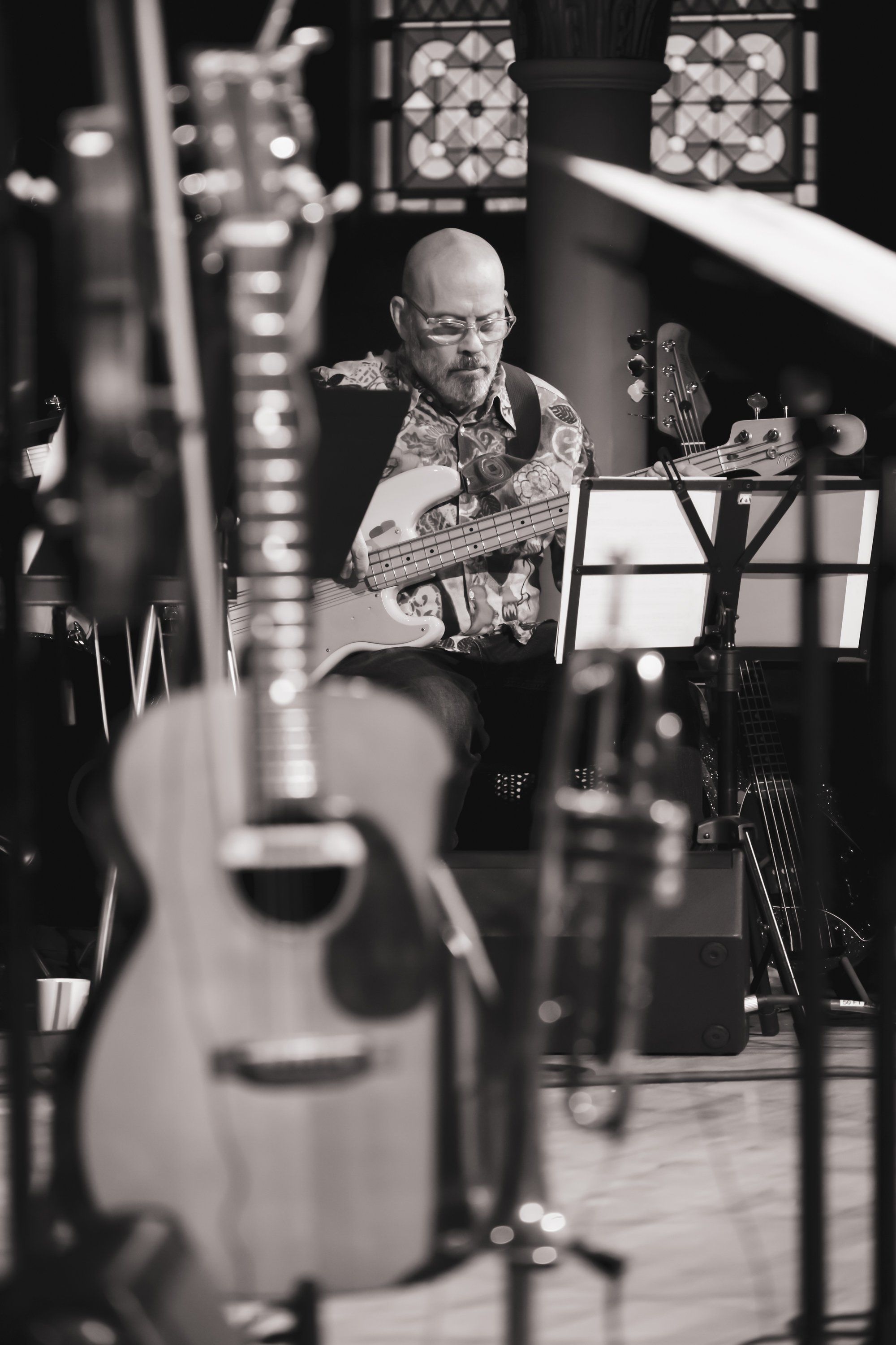 Musician in glasses performs with acoustic guitar on concert stage, surrounded by warm stage lighting and musical instruments in soft focus