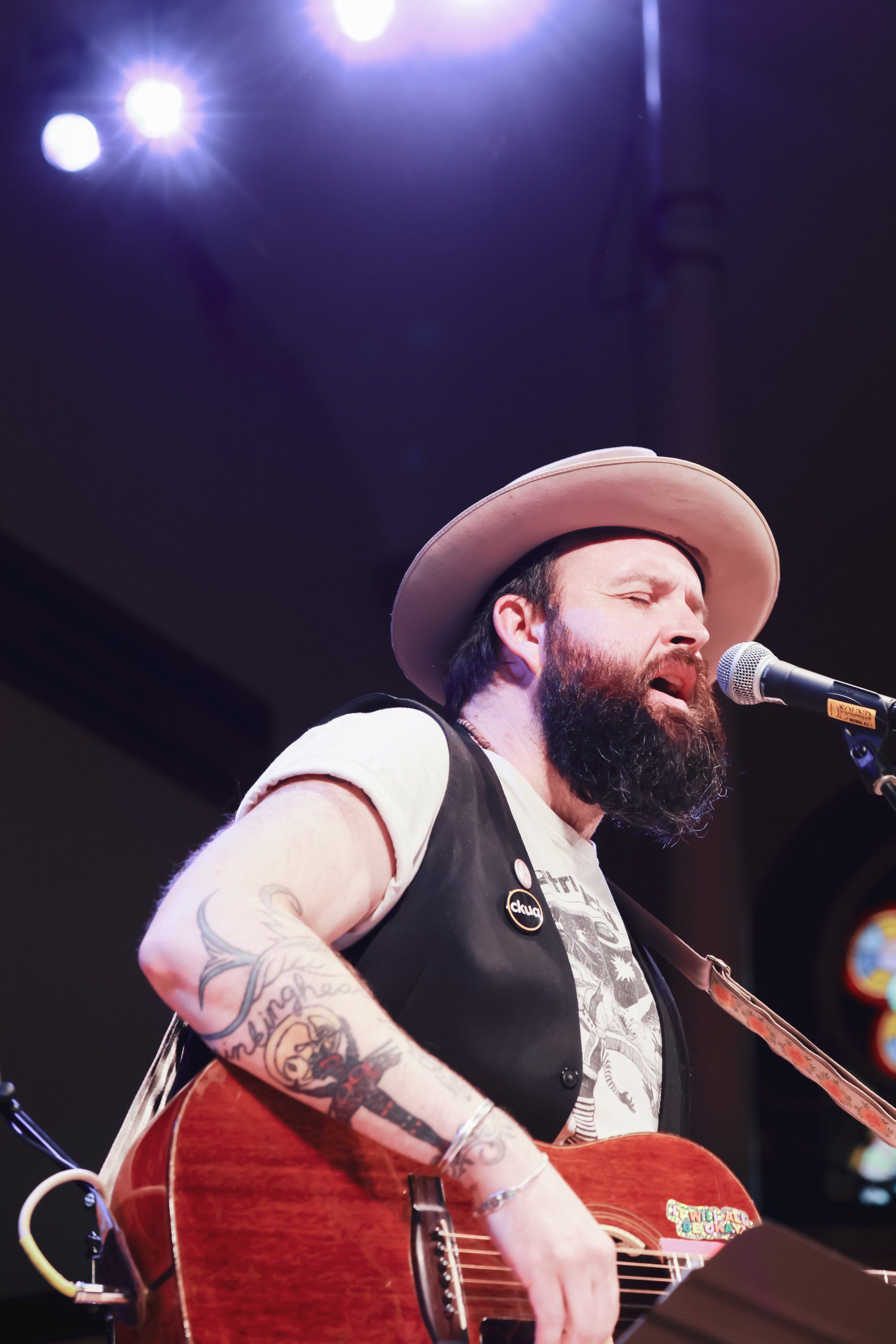 Bearded musician in pink cowboy hat and red guitar performs under stage lights, singing passionately into microphone