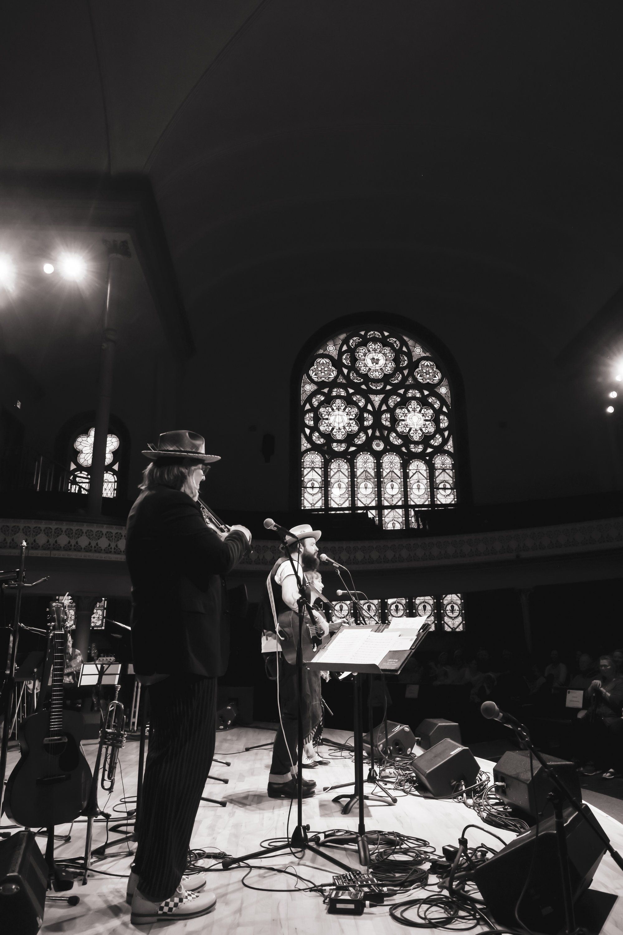 Musicians perform on stage in a grand cathedral, dramatic spotlight illuminating the ornate rose window behind them in black and white