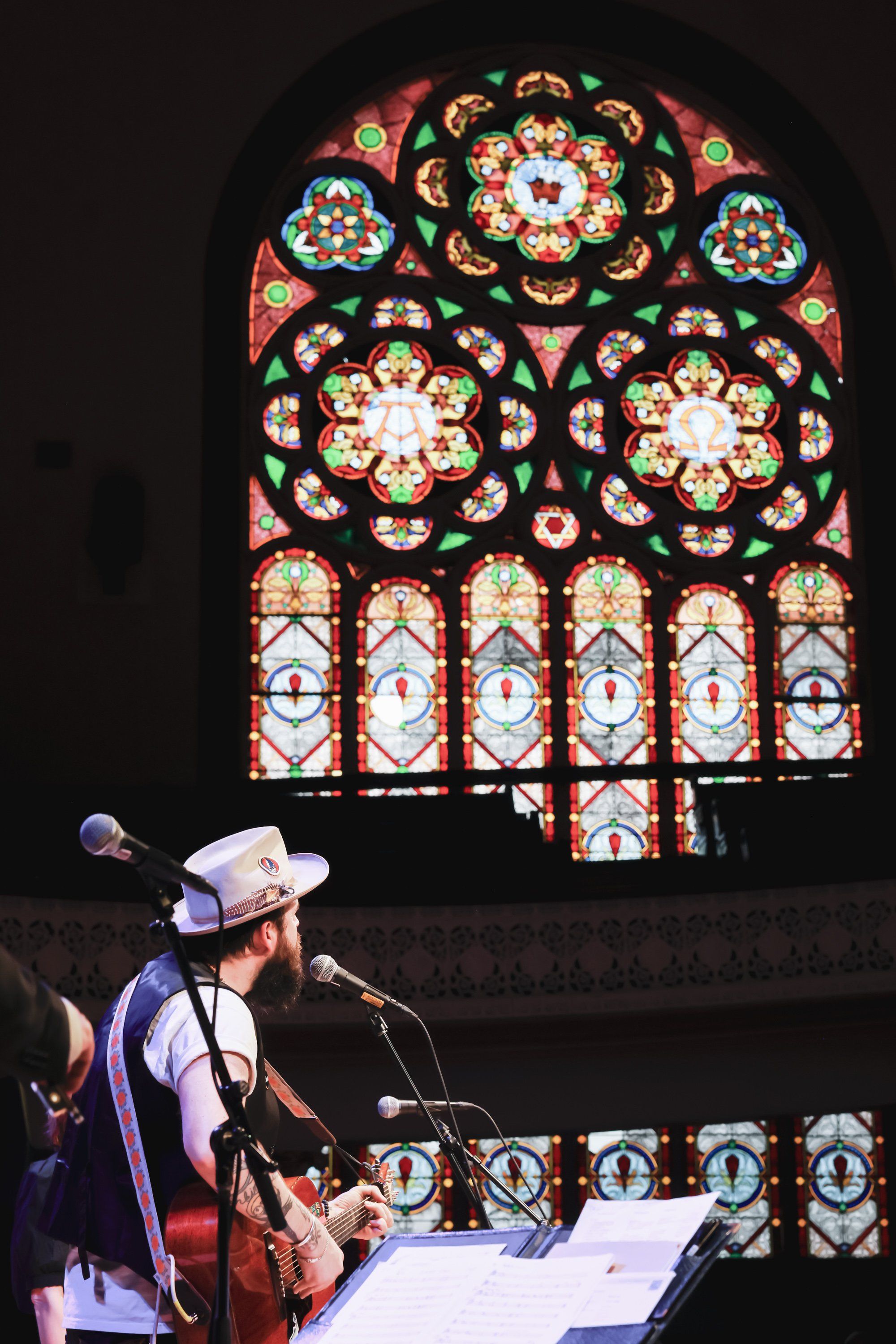 Musician in cowboy hat performs at microphone before ornate stained glass window in candlelit cathedral setting