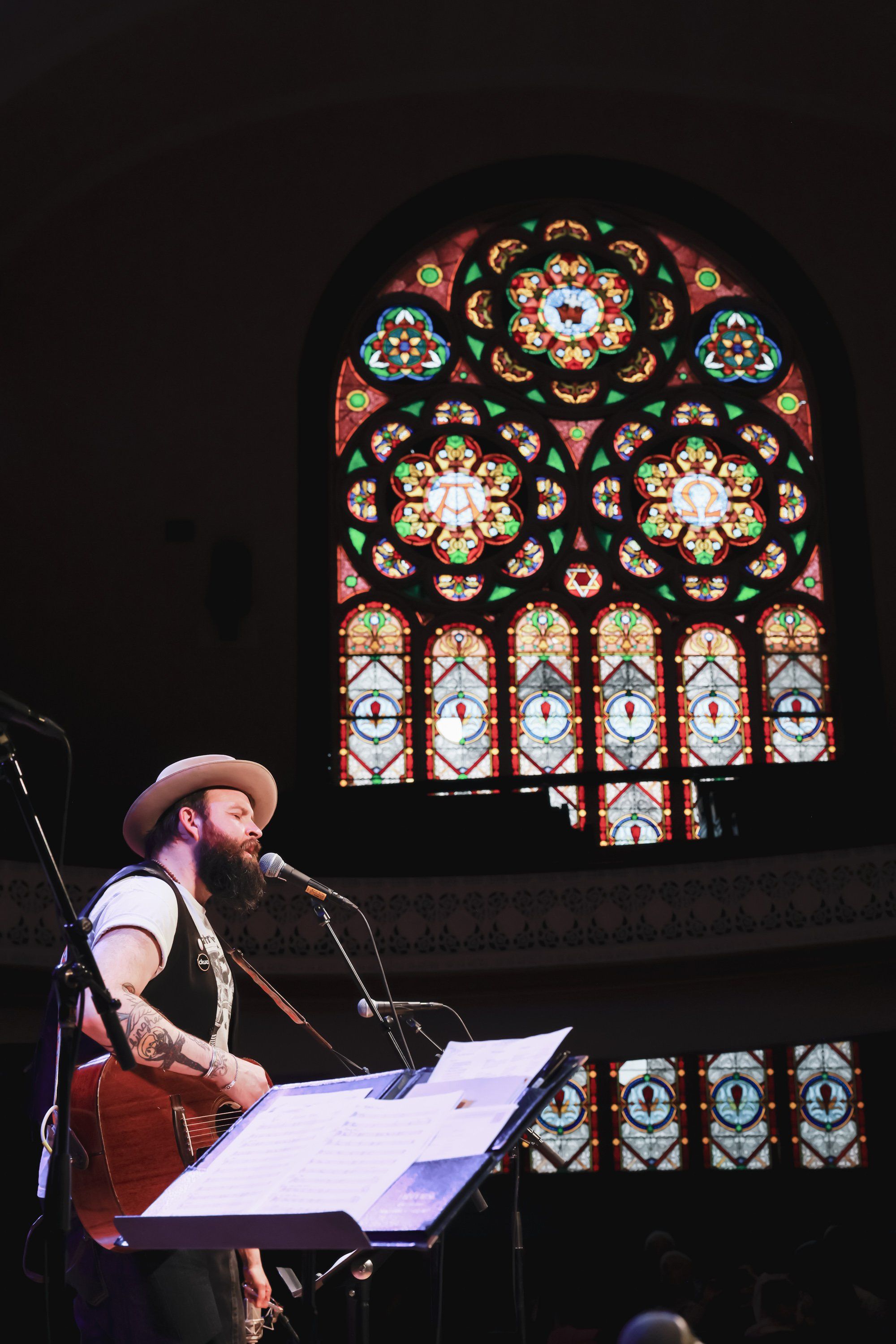 Bearded musician with guitar performing at microphone against ornate stained-glass window in candlelit cathedral setting