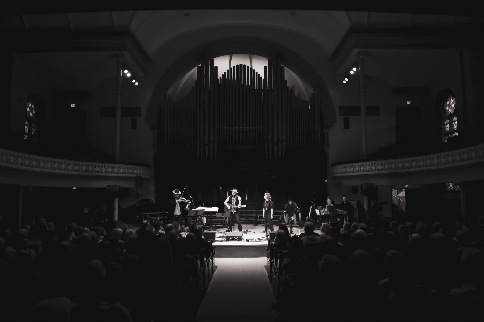 Musicians perform on a spotlit stage beneath a grand organ in a darkened concert hall filled with seated audience members