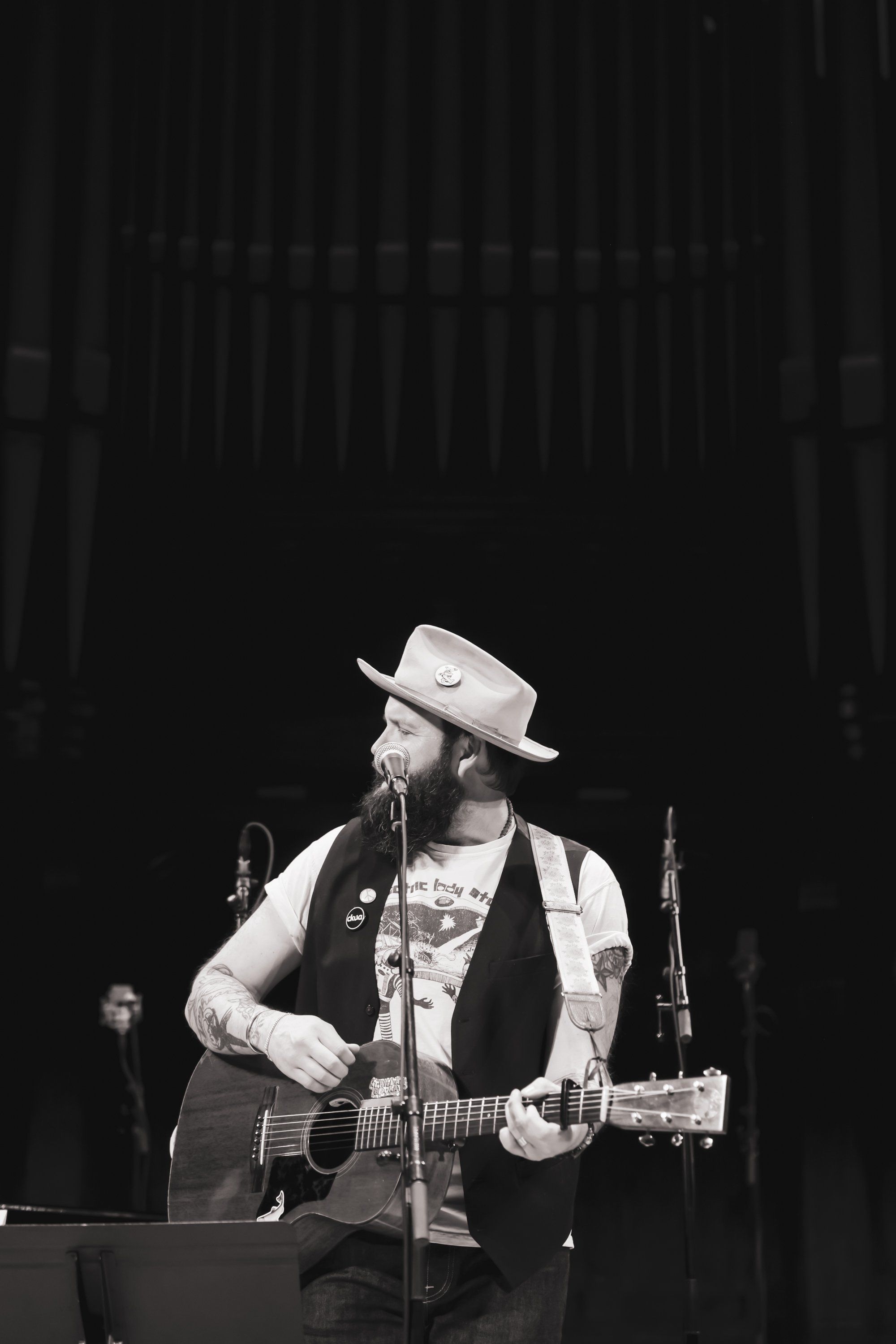 Bearded musician in cowboy hat and vest plays acoustic guitar under stage lights during an intimate concert performance