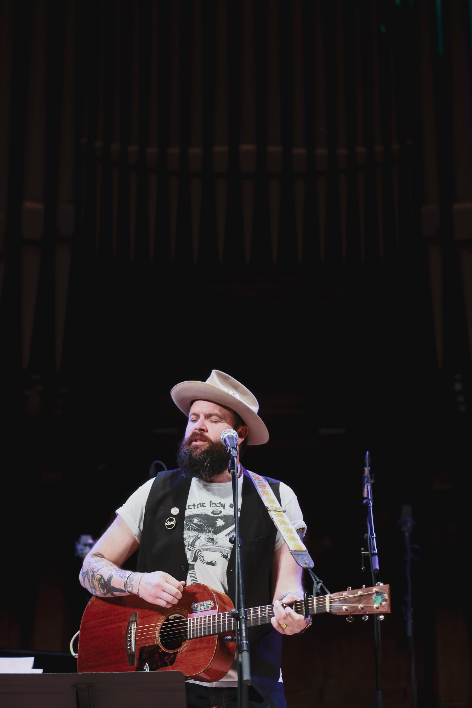 Bearded musician in cowboy hat and red acoustic guitar performs on stage against dark backdrop, spotlight illuminating his focused expression