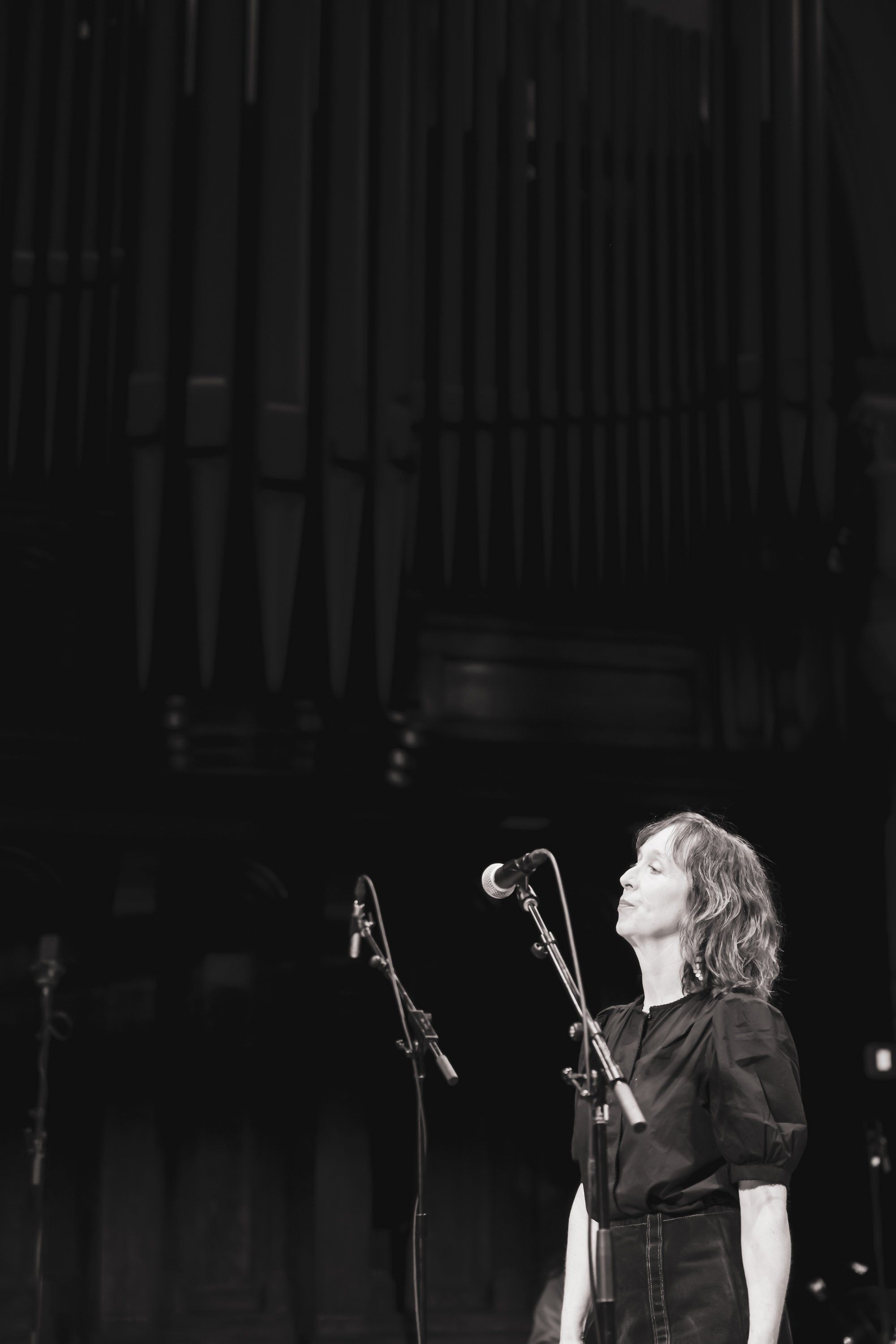 Woman with blonde wavy hair singing at microphone in dramatic black and white stage lighting, wearing dark clothing against moody architectural background