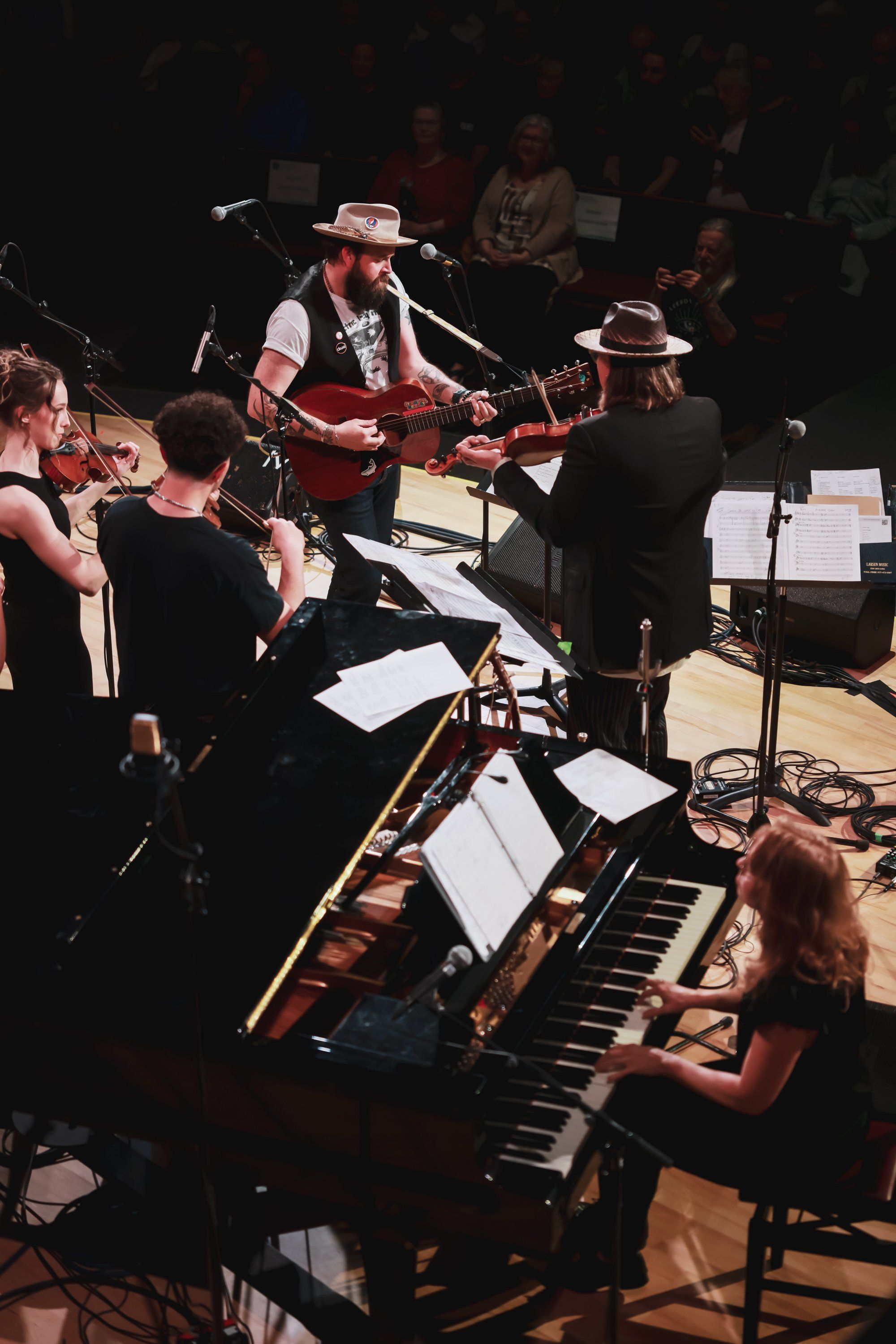Live concert performance with musicians in black clothing playing keyboards and guitar under warm stage lighting, sheet music visible on stands