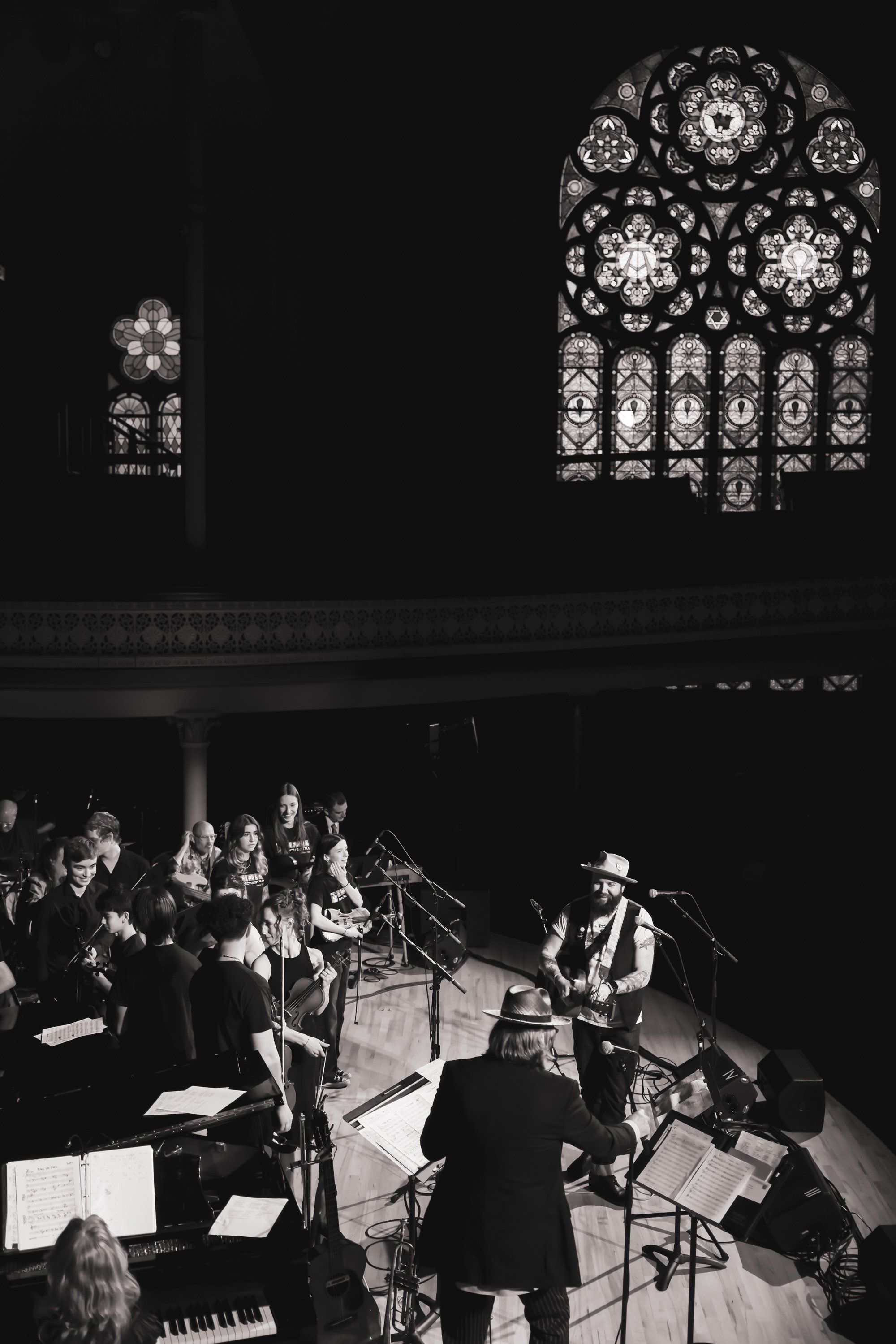 Musicians perform under Gothic stained glass windows in a stone chapel, lit dramatically in black and white