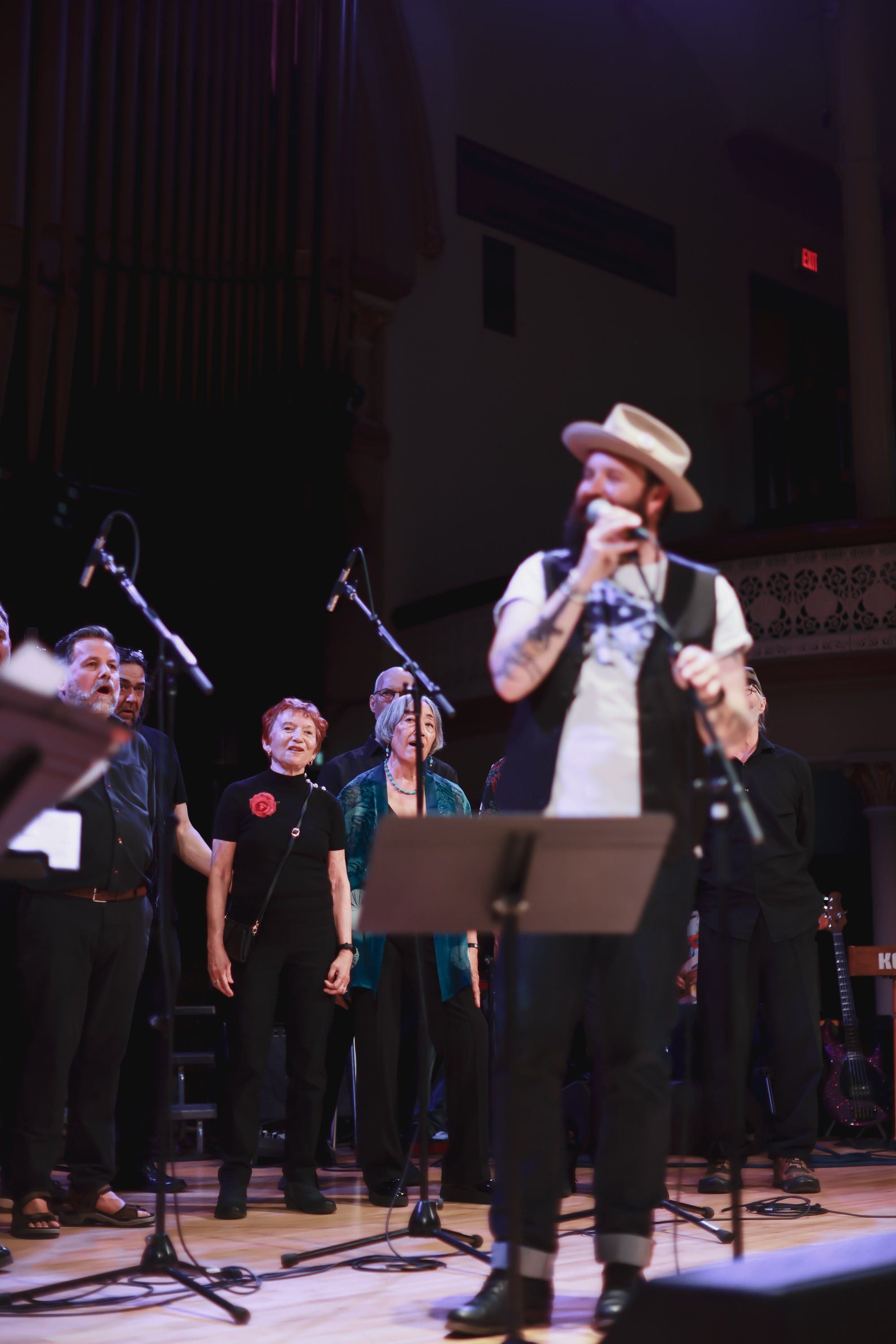 Daniel Lapp performs at center stage in a cream-colored hat and vest, flanked by backup singers at microphones in a warmly-lit concert hall