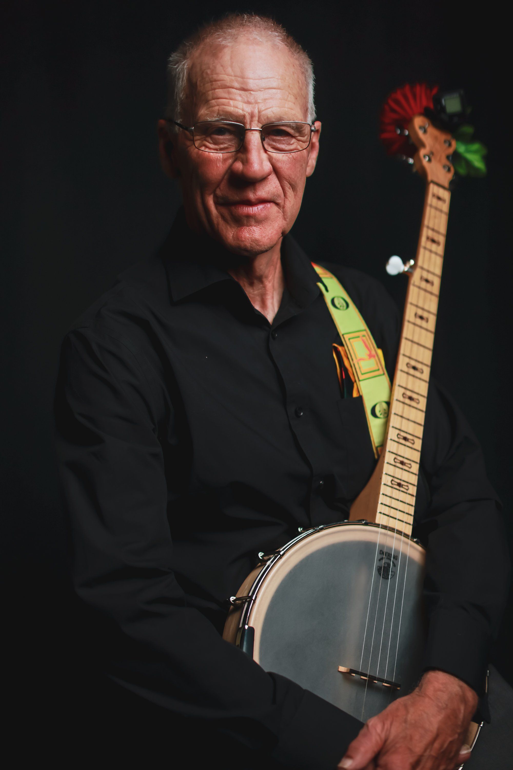 Older man with glasses holding a banjo against dark background, wearing black shirt and colorful strap, direct gaze at camera