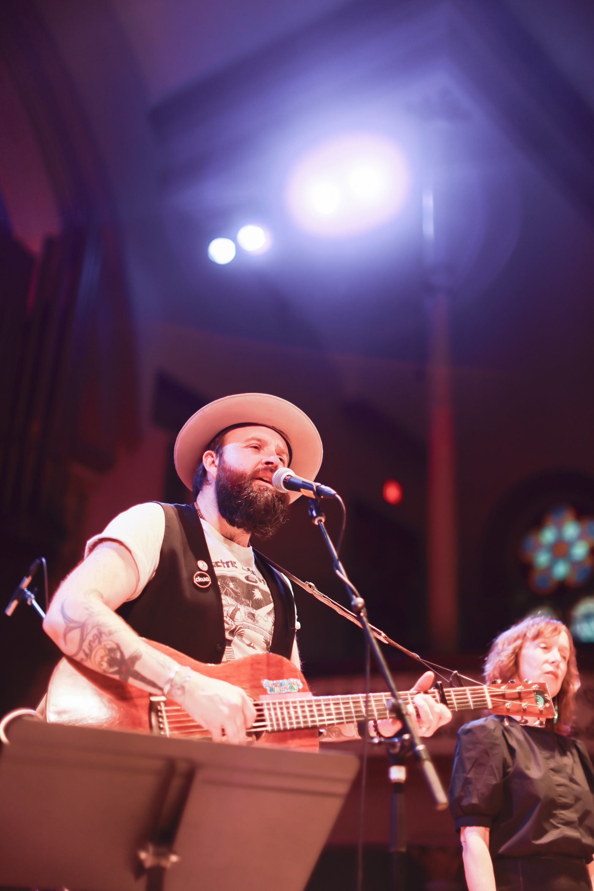 Bearded musician in cowboy hat and vest performing with acoustic guitar under warm stage lights at an indoor concert venue