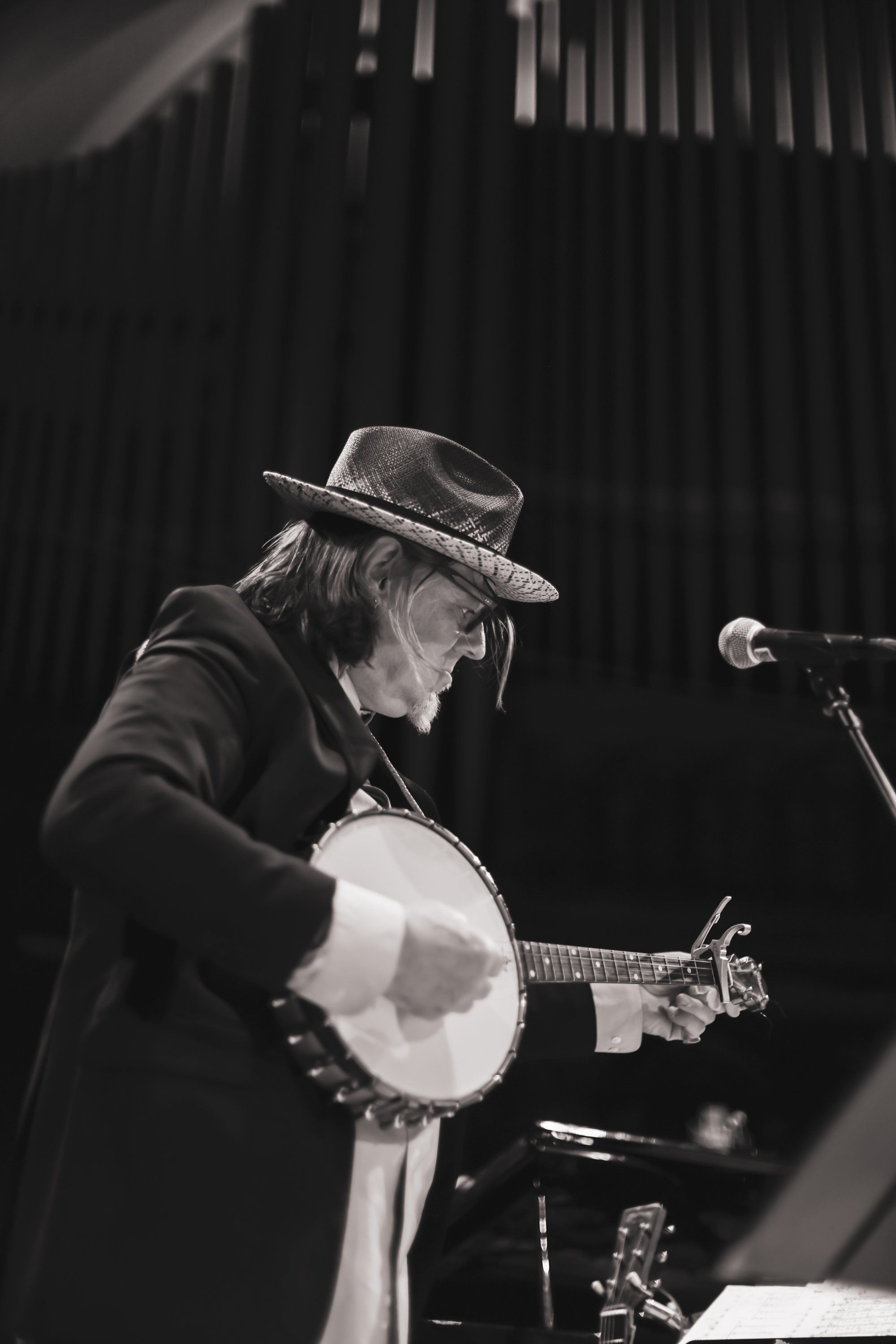 Musician in cowboy hat playing banjo at microphone, dramatic stage lighting casting shadows on dark backdrop