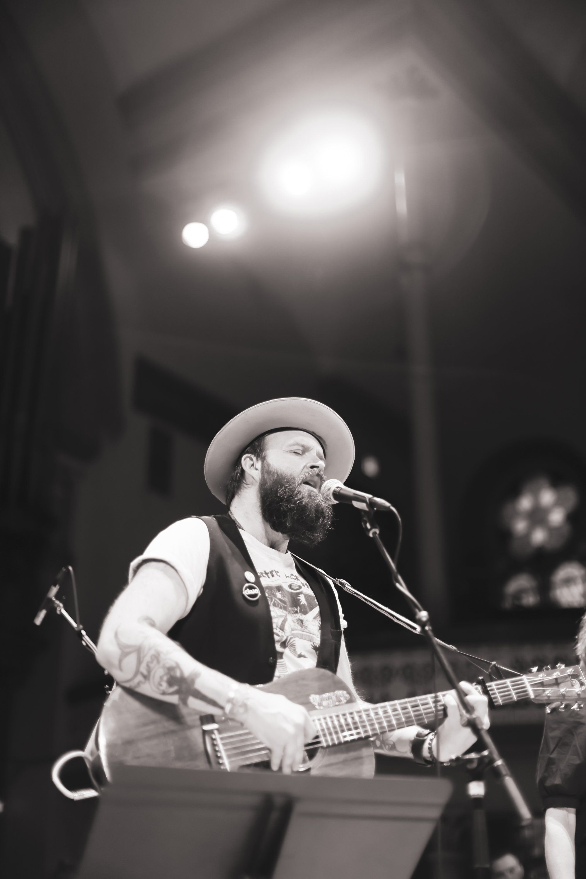 Bearded musician in cowboy hat and vest playing acoustic guitar at microphone under stage lights, black and white concert photography