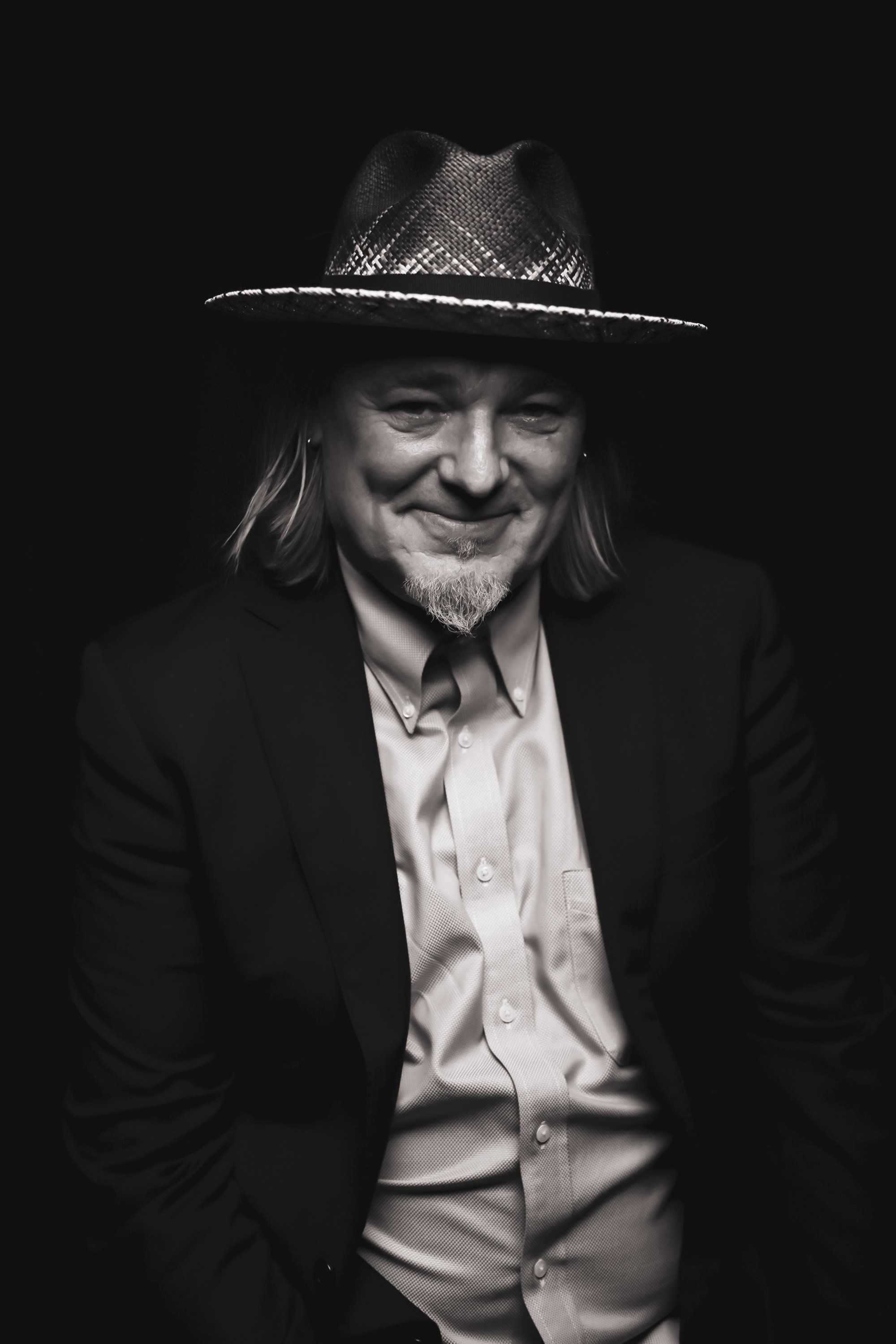 Man in cowboy hat and dark blazer smiles warmly against black background, lit dramatically from the side