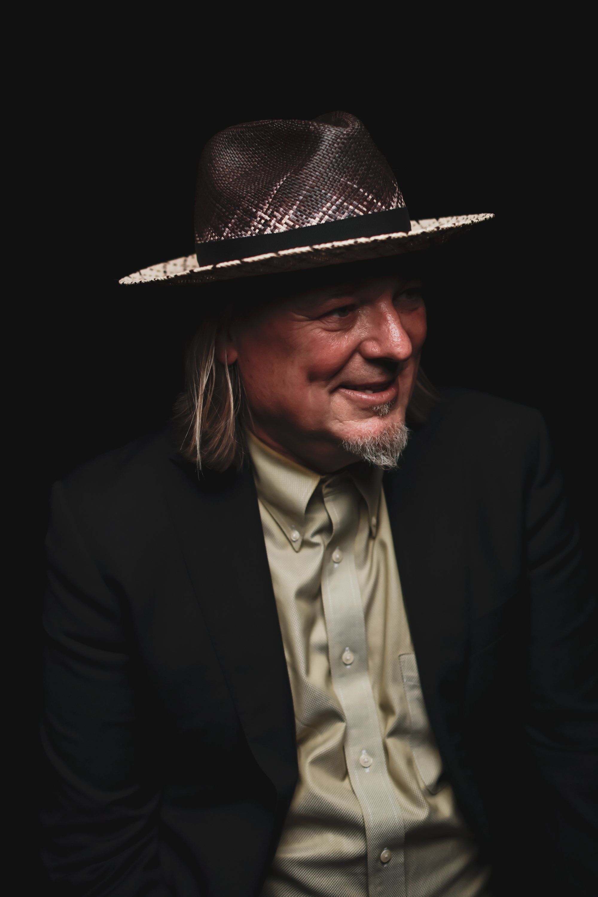 Man in black blazer and cream shirt wearing a decorative cowboy hat, smiling against dark background
