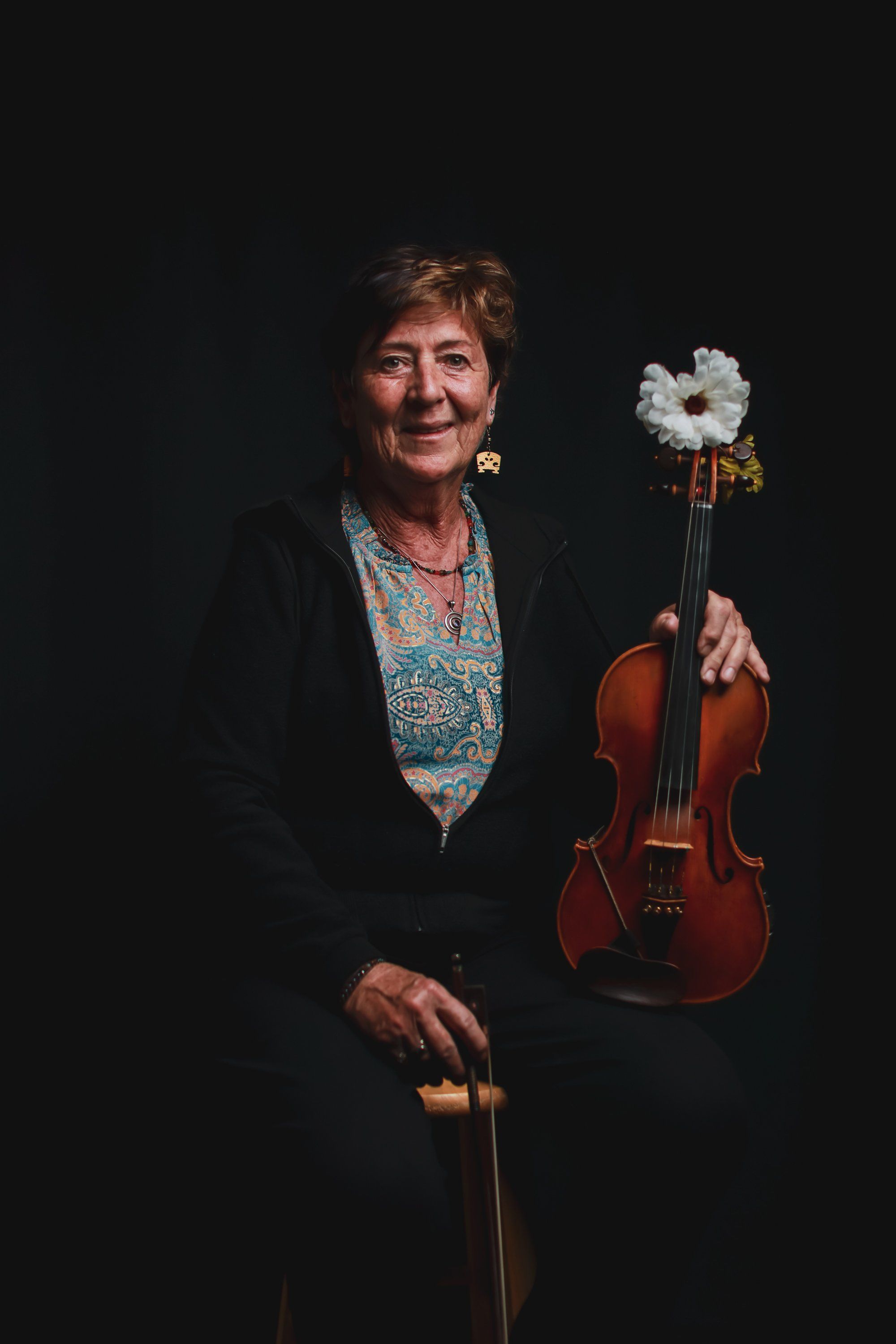 Smiling violinist holding her instrument with a white flower adorned bow against a dark background, wearing a colorful patterned blouse and dark blazer