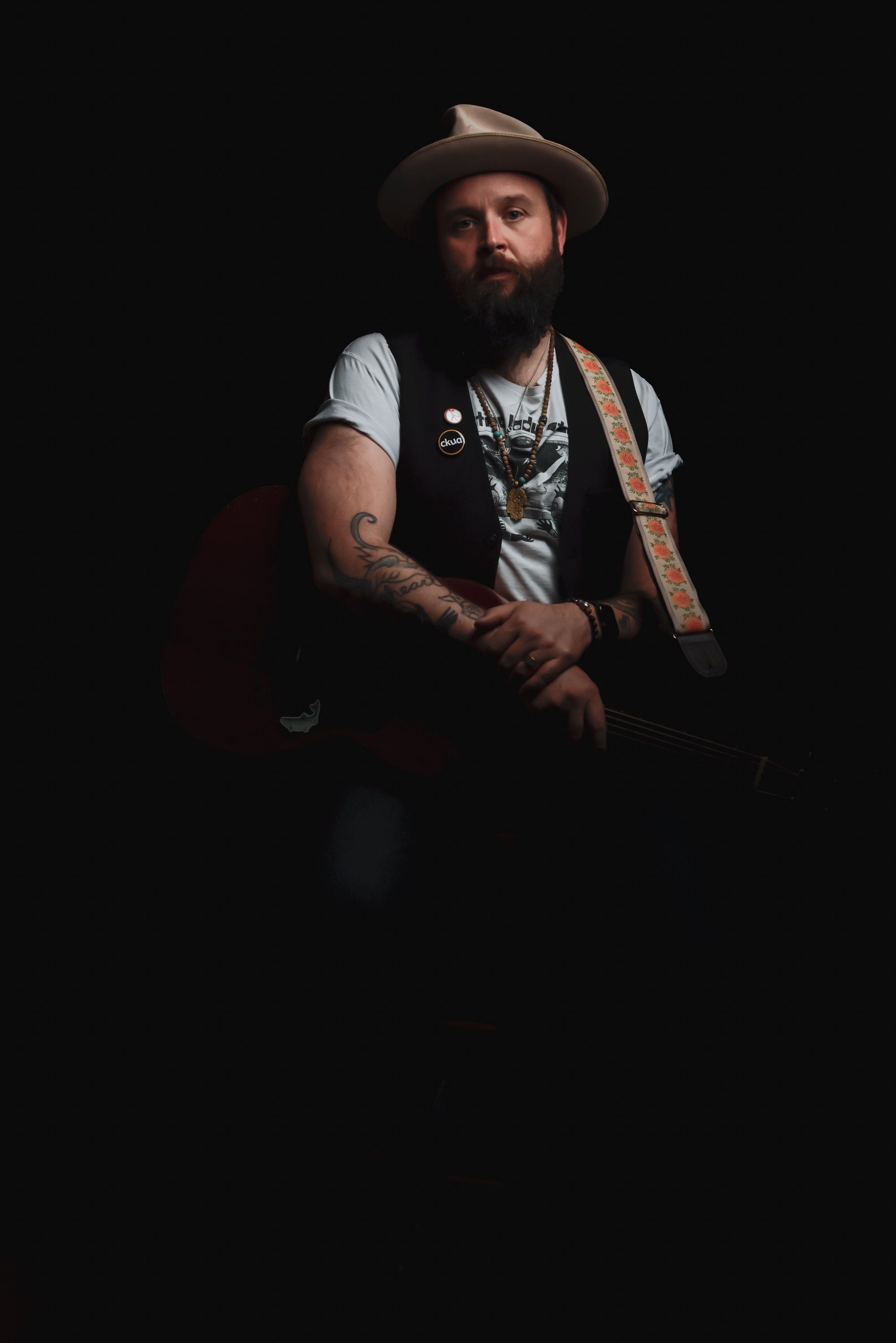 Bearded musician in cowboy hat and vest with guitar against dark background, warm studio lighting highlighting his contemplative expression