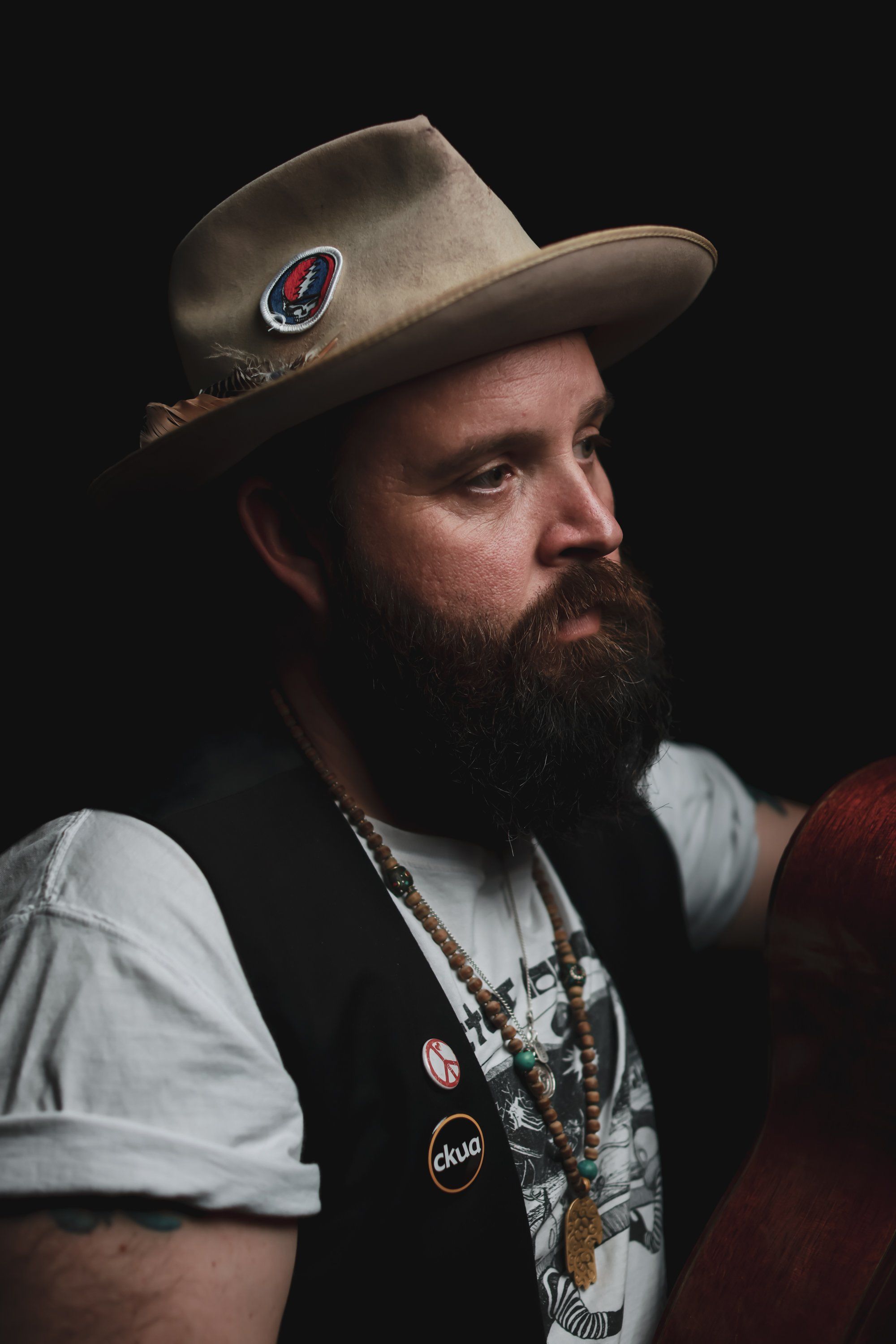 Bearded man in tan cowboy hat and layered necklaces gazes sideways against dark background, dramatic side lighting