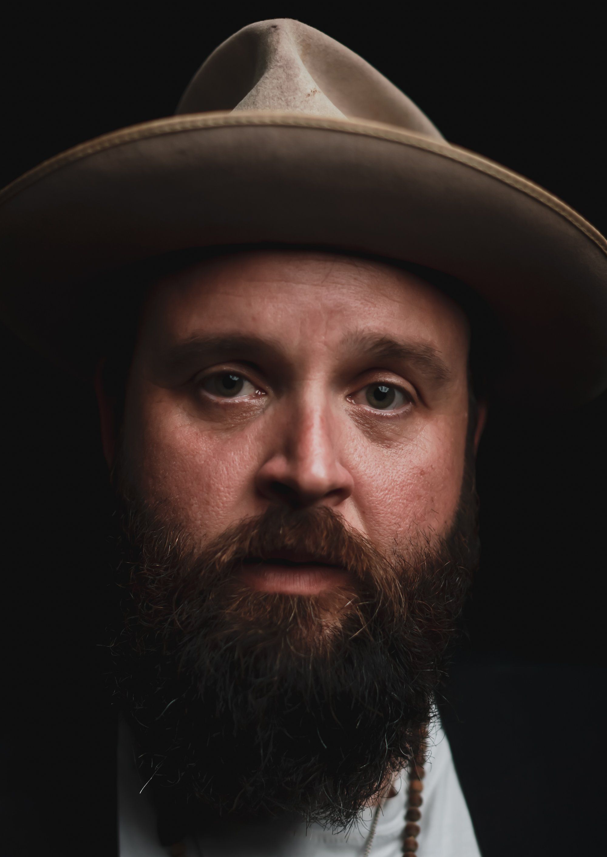 Bearded man in cowboy hat against dark background, warm studio lighting highlighting weathered features and contemplative expression