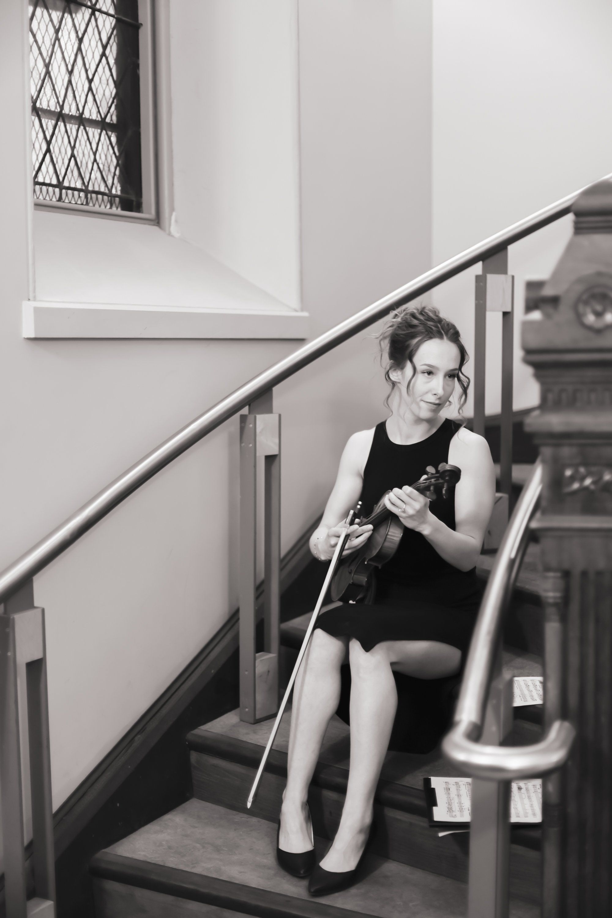 Woman sitting on staircase landing in black sleeveless top, holding violin, contemplative expression in monochrome photograph