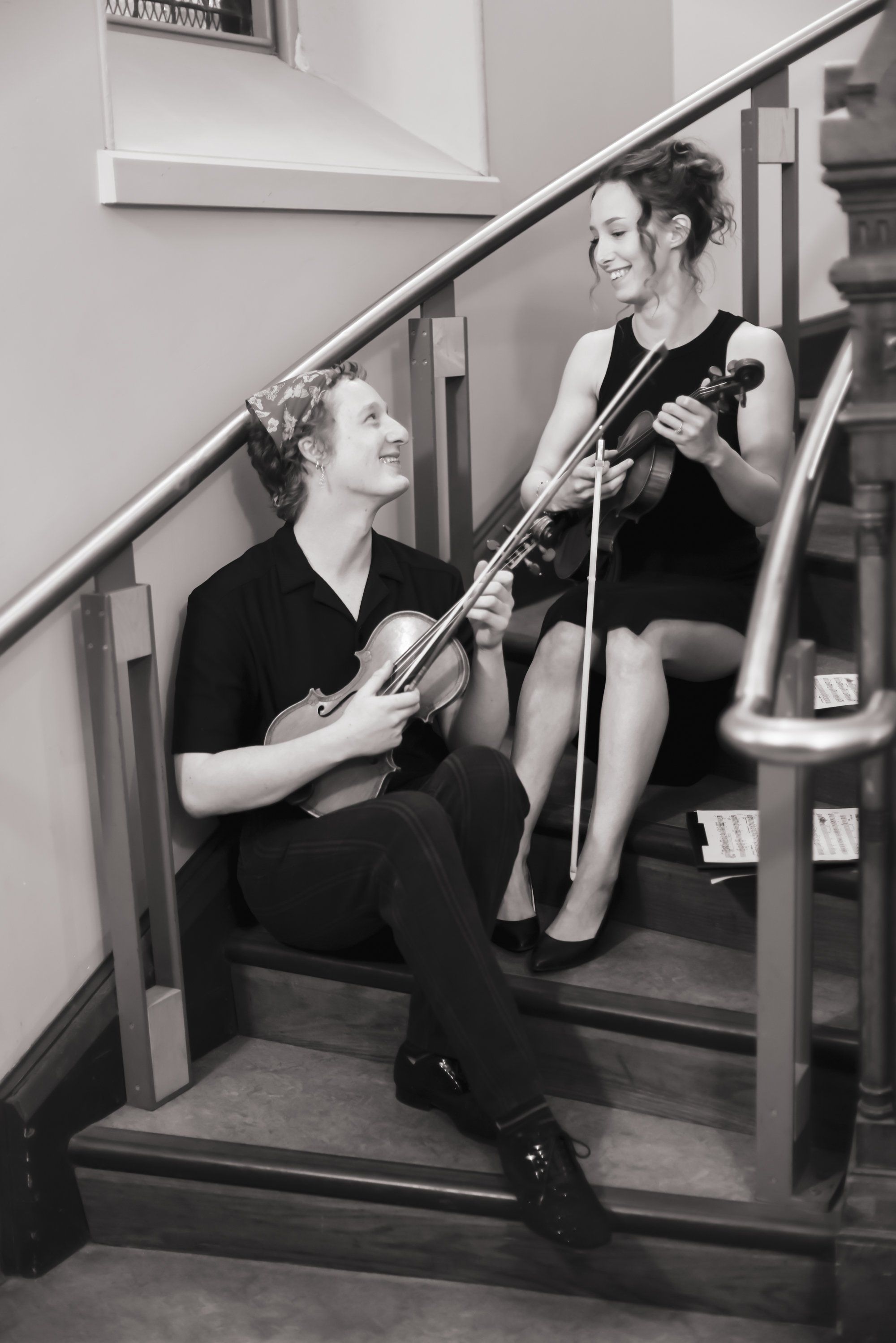 Two musicians sit on interior stairs in black clothing, one holding a violin and one a cello, gazing upward in a black and white portrait