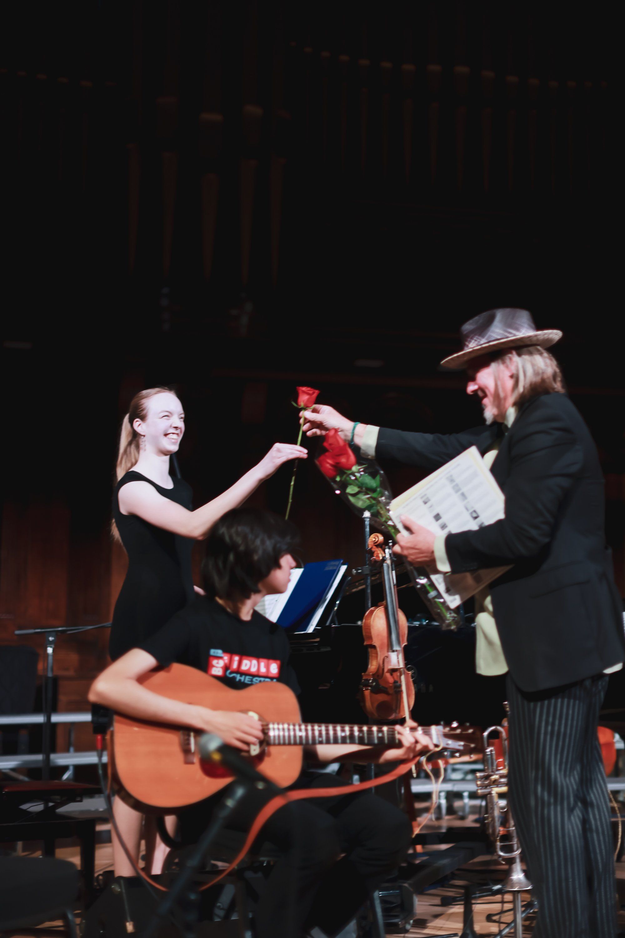 Musician in blue shirt plays acoustic guitar while woman in black dress receives red roses from man in hat and suit on dimly lit stage